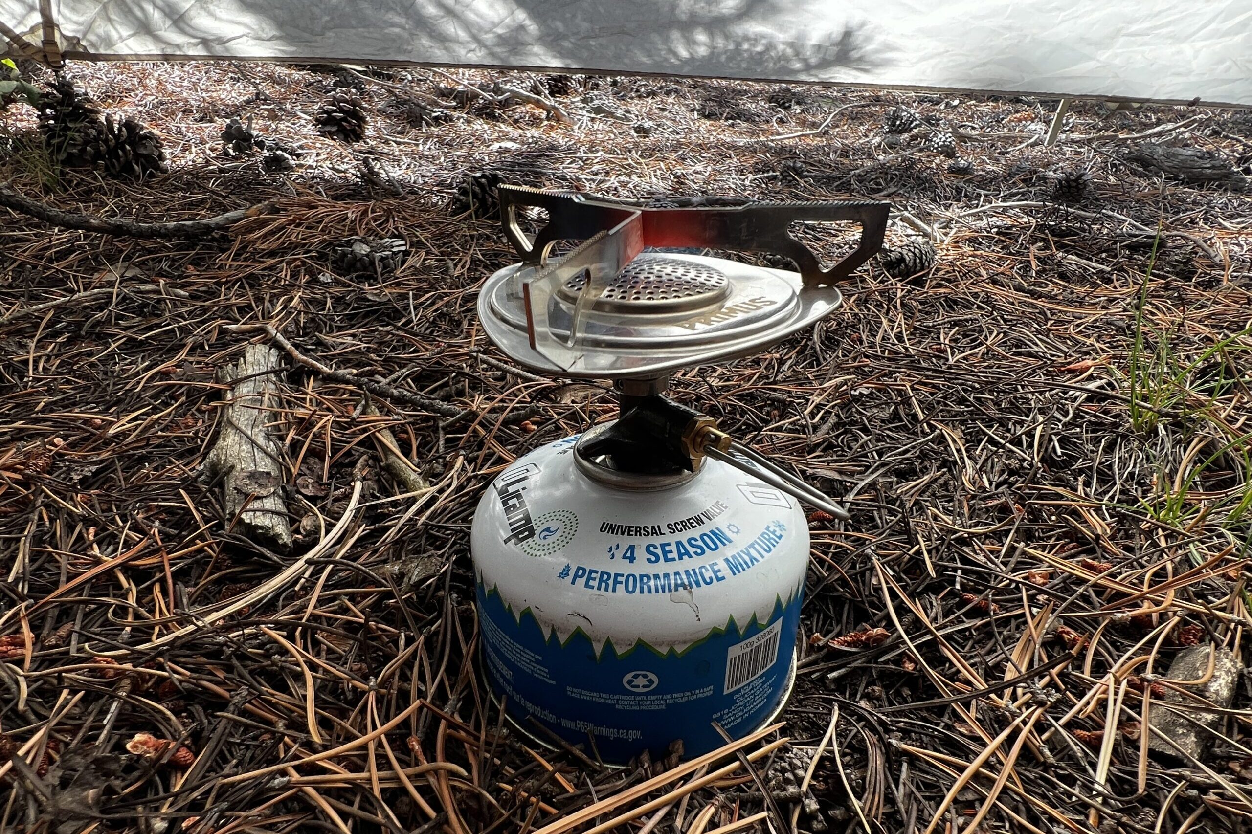 The Primus Essential Trail is screwed onto a fuel canister and its flame is active. The center of the pot support is red hot. A tent wall and pine needles are in the background.