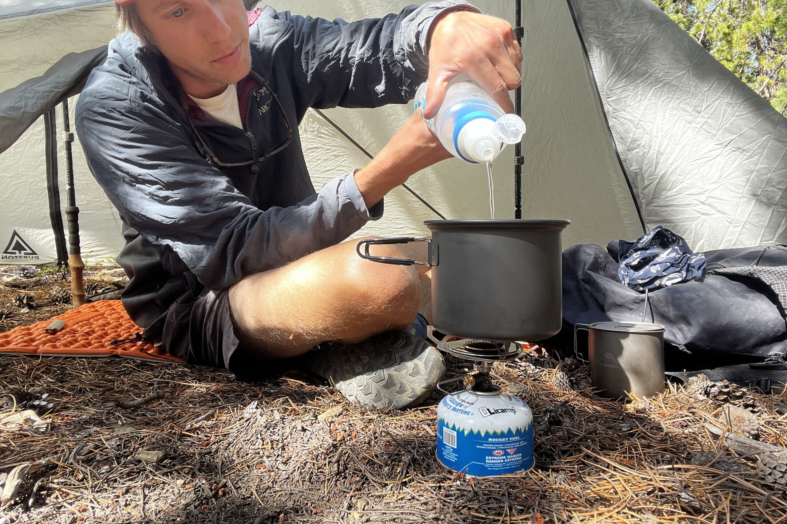 A hiker filters water into a pot that is sitting on the primus essential trail stove. Tent, backpack, and other gear are in the background.