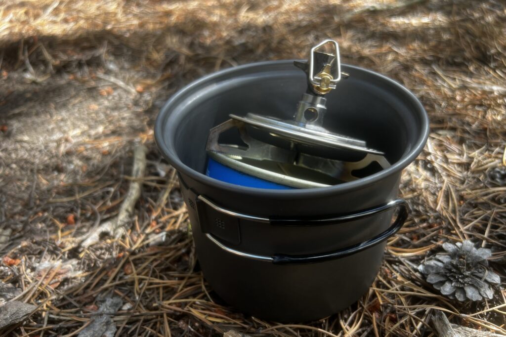 The Primus Essential stove packed into a one liter pot with an 8oz fuel canister. The stove is sticking out of the pot just a little. The whole thing sites on a bed of pine needles.