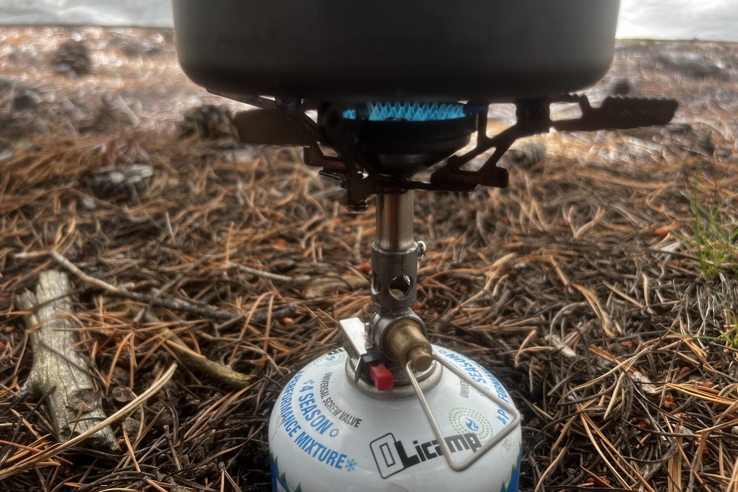 A close up of a pot sitting on the flame and pot support of the WindMaster stove. The stove is on a fuel canister. A tent wall and pine needles are in the background.