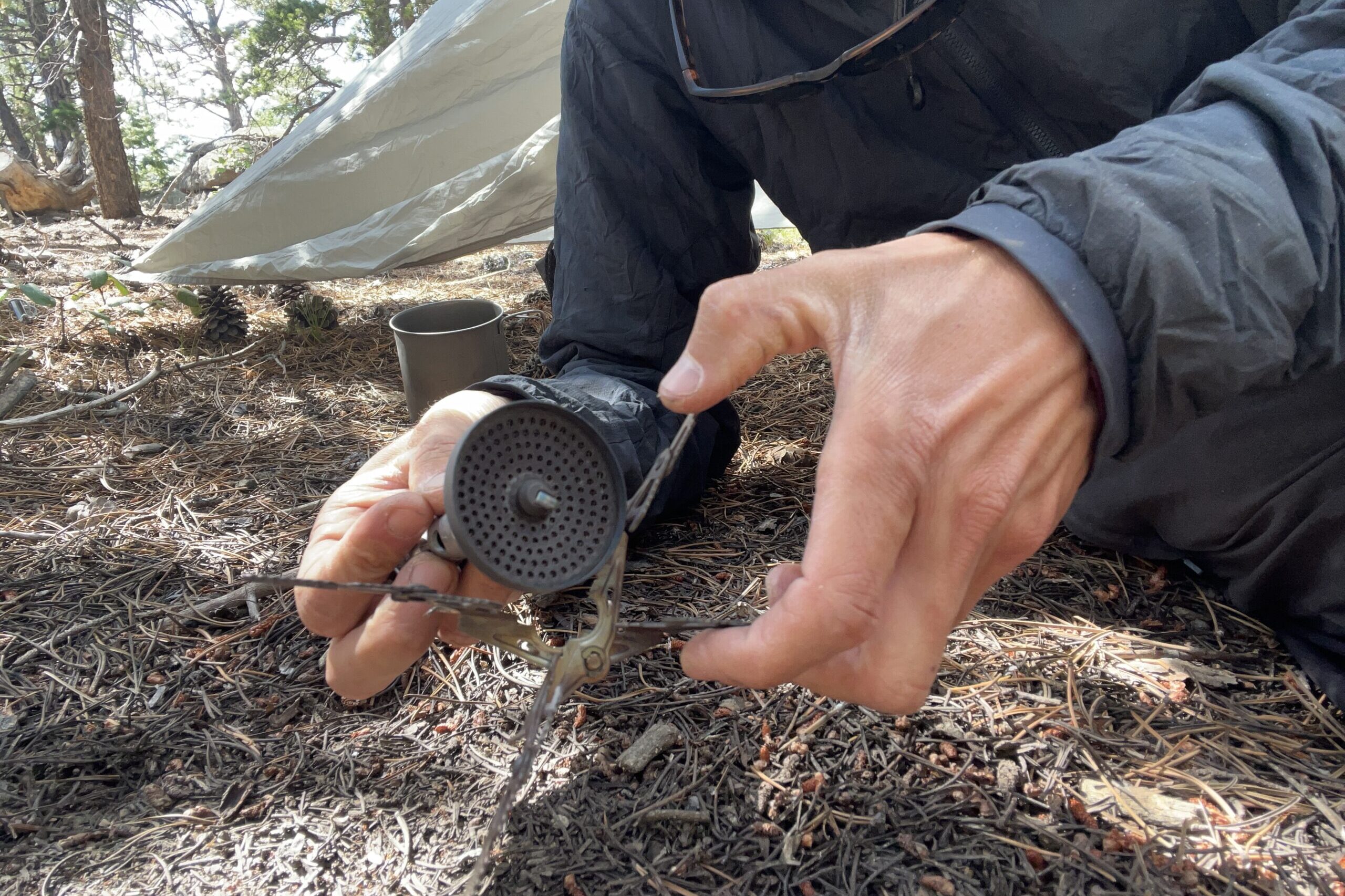 A hiker leans towards the camera and demonstrates the installing of the 4Flex support on the WIndMaster stove. Part of a tent, a pot and trees are in the background.