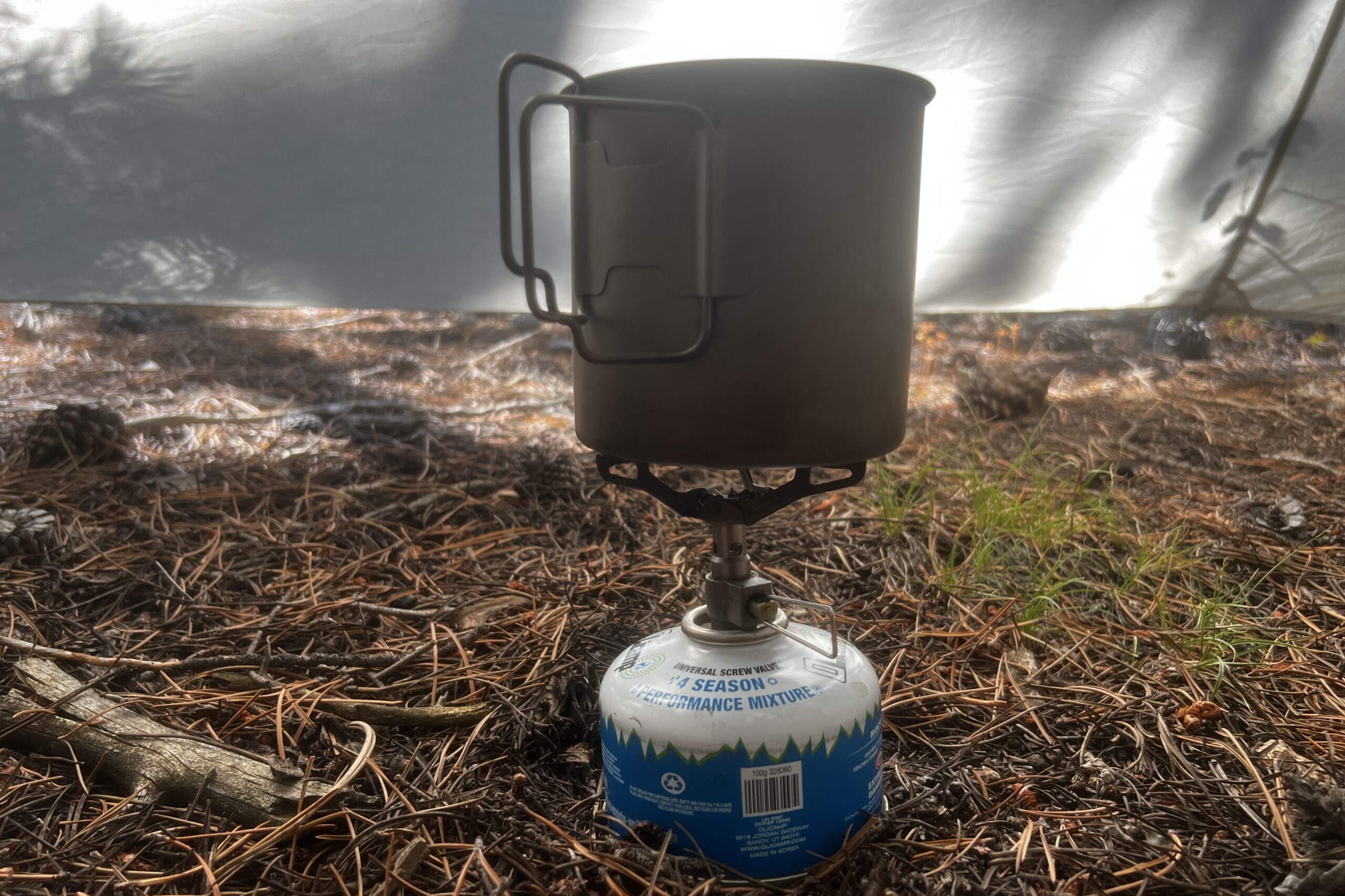 A small pot sitting on the BRS 3000T and fuel canister with no flame. The background is a tent wall and pine needle floor.