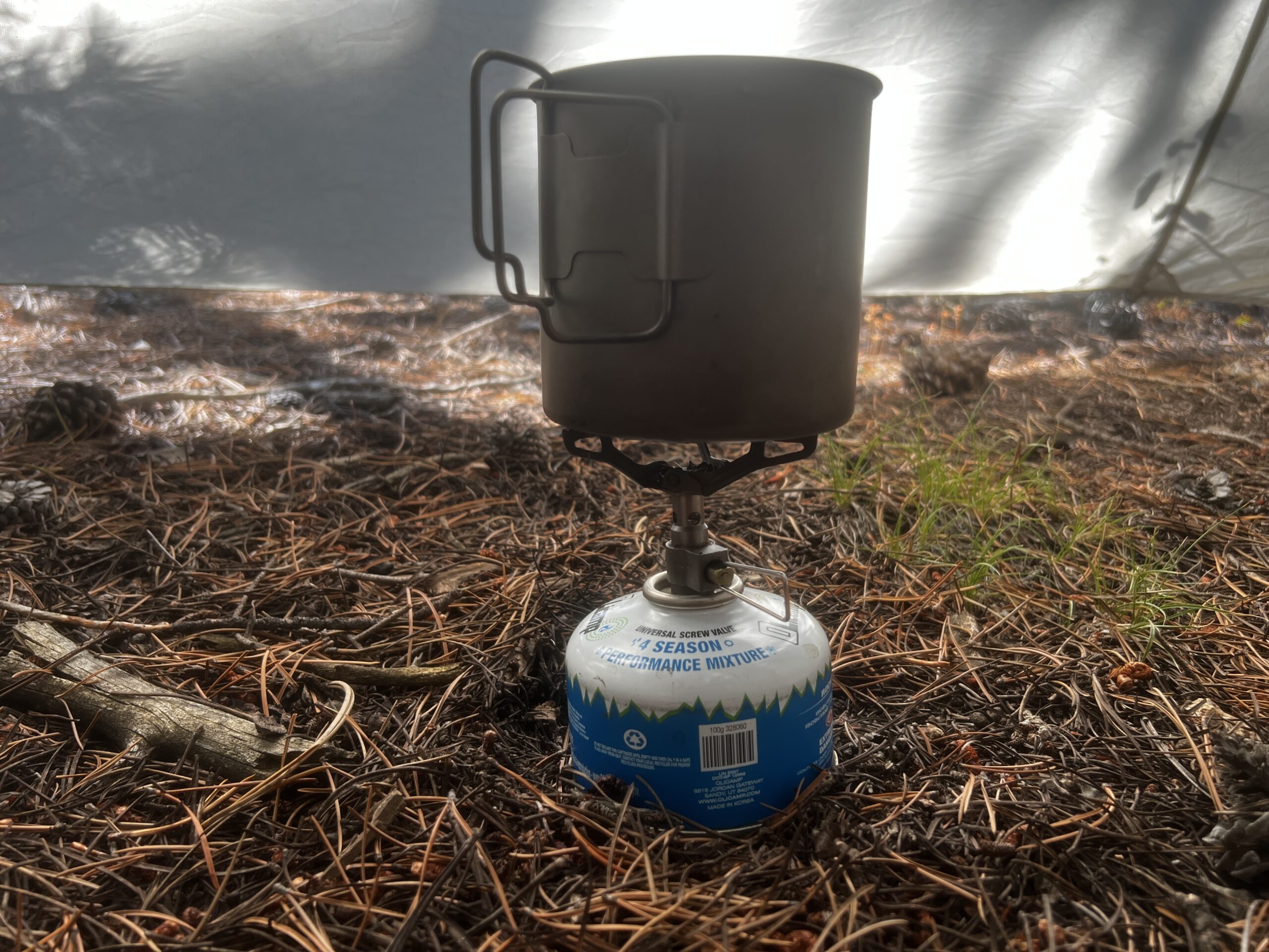 A small pot sitting on the BRS 3000T and fuel canister with no flame. The background is a tent wall and pine needle floor.