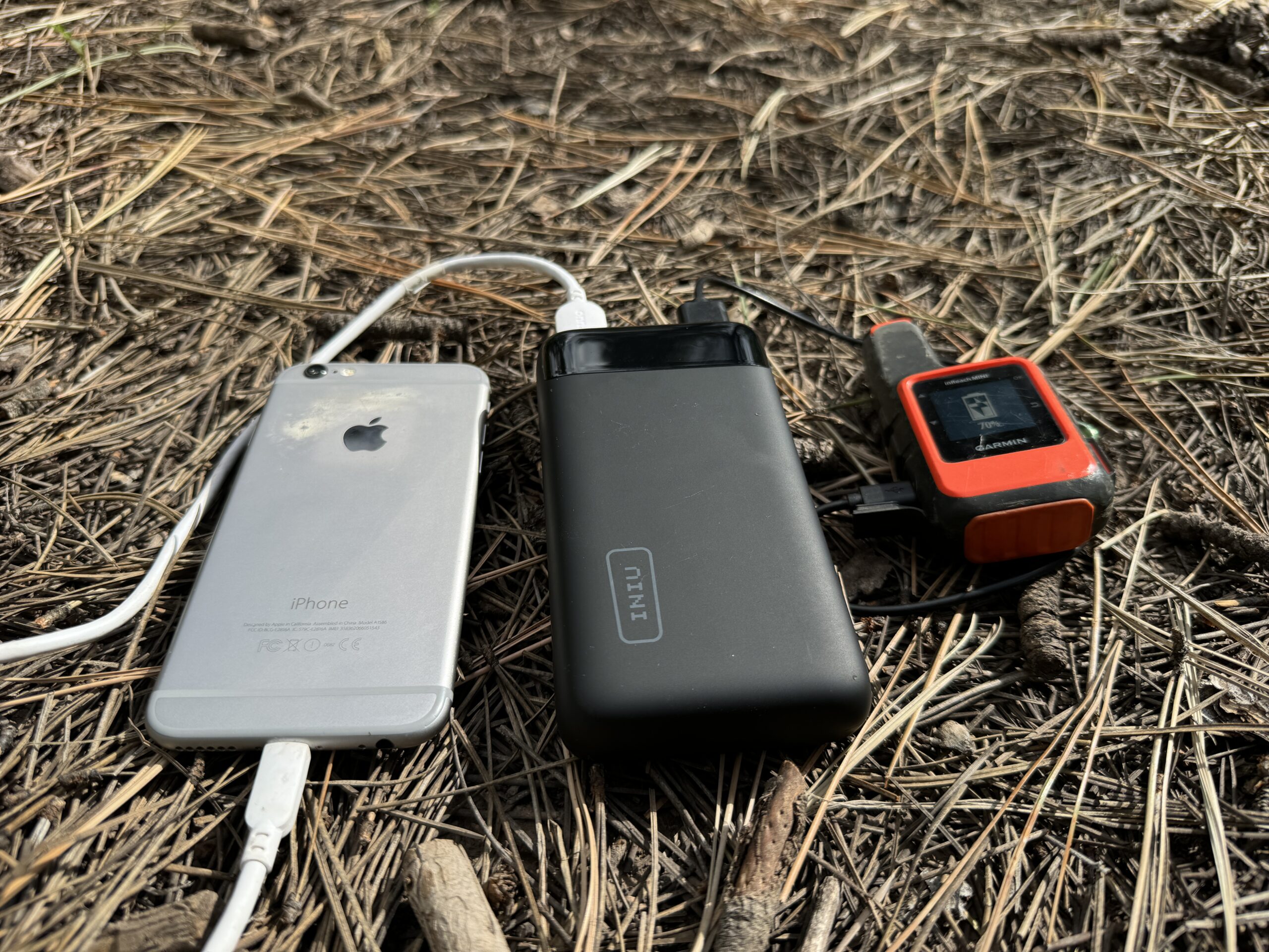 Close up of INIU 20000 PD power bank charging a phone and the Garmin Inreach while sitting on some pine needles.