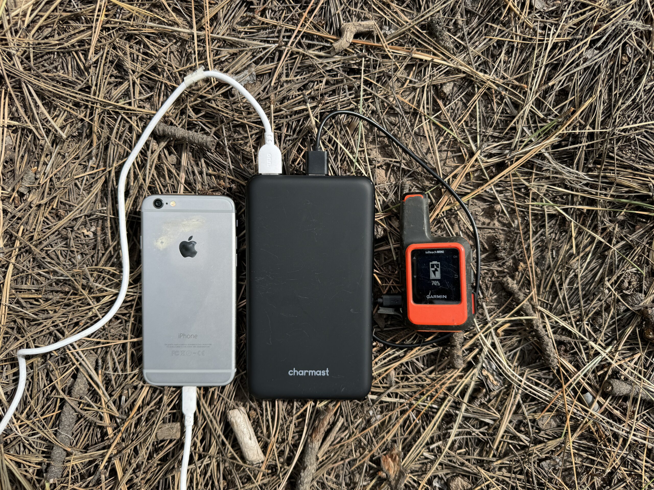 Close up of the Charmast power bank charging a phone and the Garmin Inreach while sitting on some pine needles.