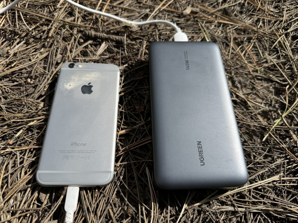 Close up of the UGREEN power bank charging a phone and the Garmin Inreach while sitting on some pine needles.