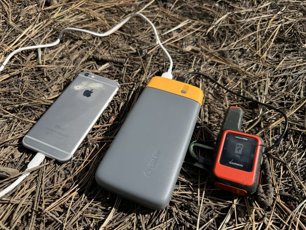 Close up of the BioLite power bank charging a phone and the Garmin Inreach while sitting on some pine needles.