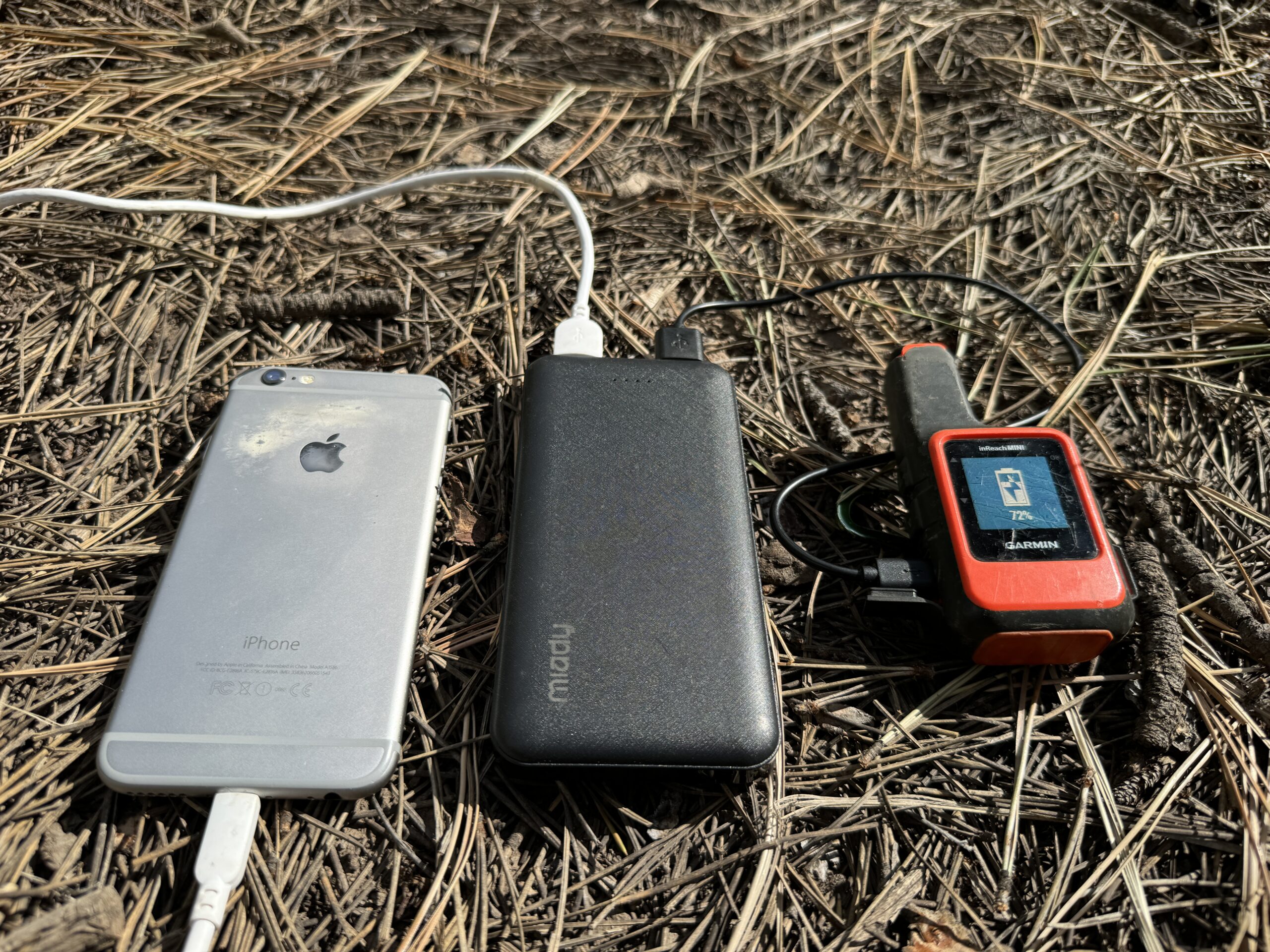 Close up of the Miady power bank charging a phone and the Garmin Inreach while sitting on some pine needles.