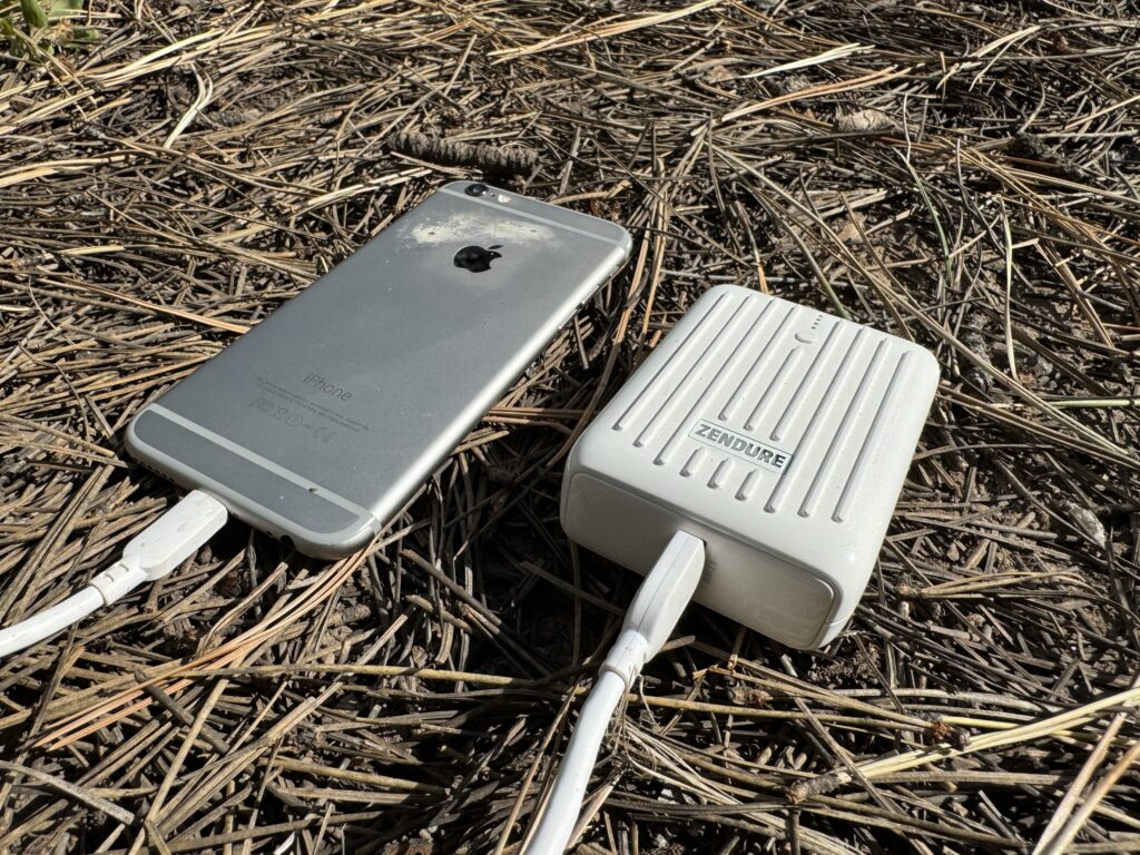 Close up of the Zendure power bank charging a phone while sitting on some pine needles.