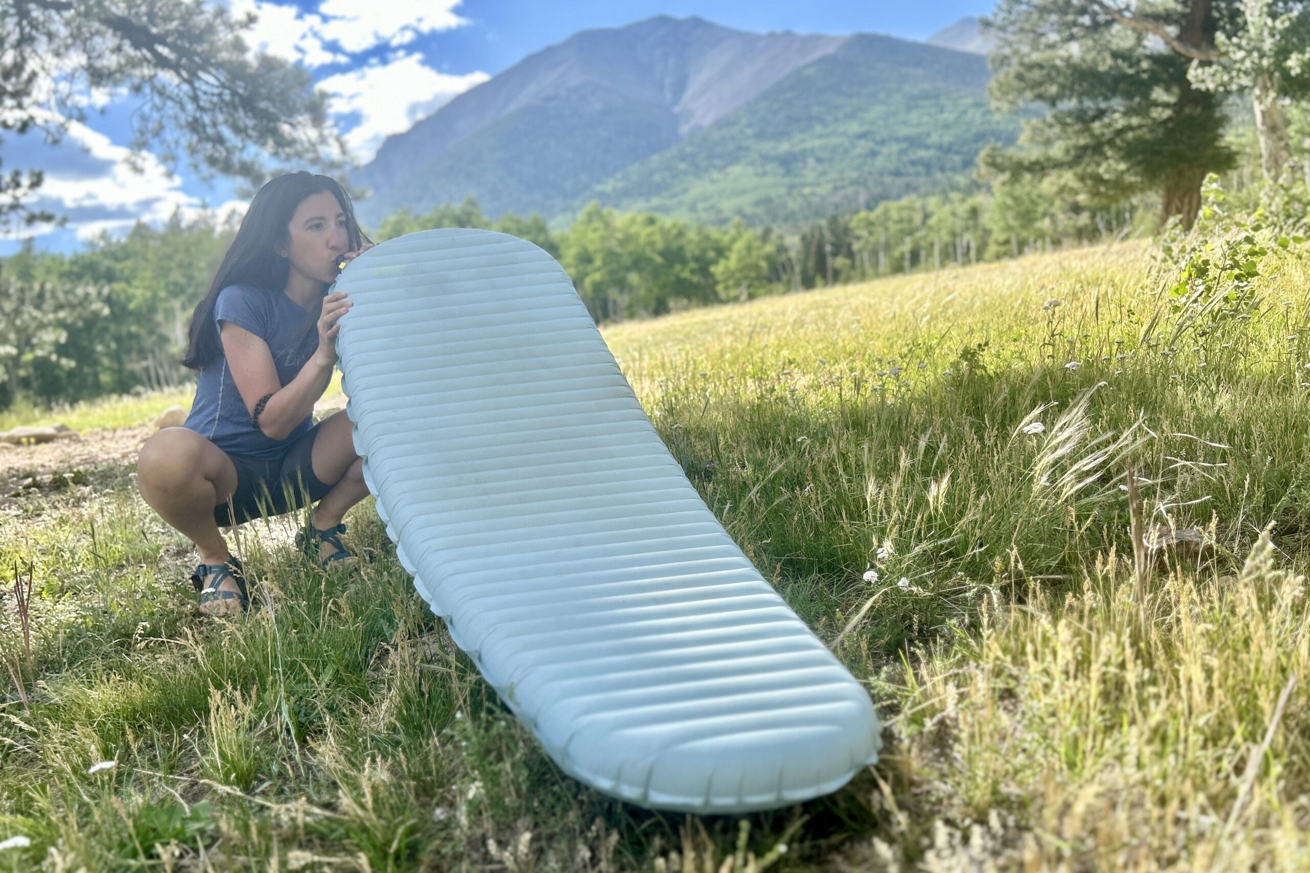 A woman blows up a sleeping pad valve with a huge field and mountain in background.