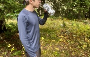 A hiker sips from a water bottle while wearing the Patagonia Capilene base layer. Trees are in the background.