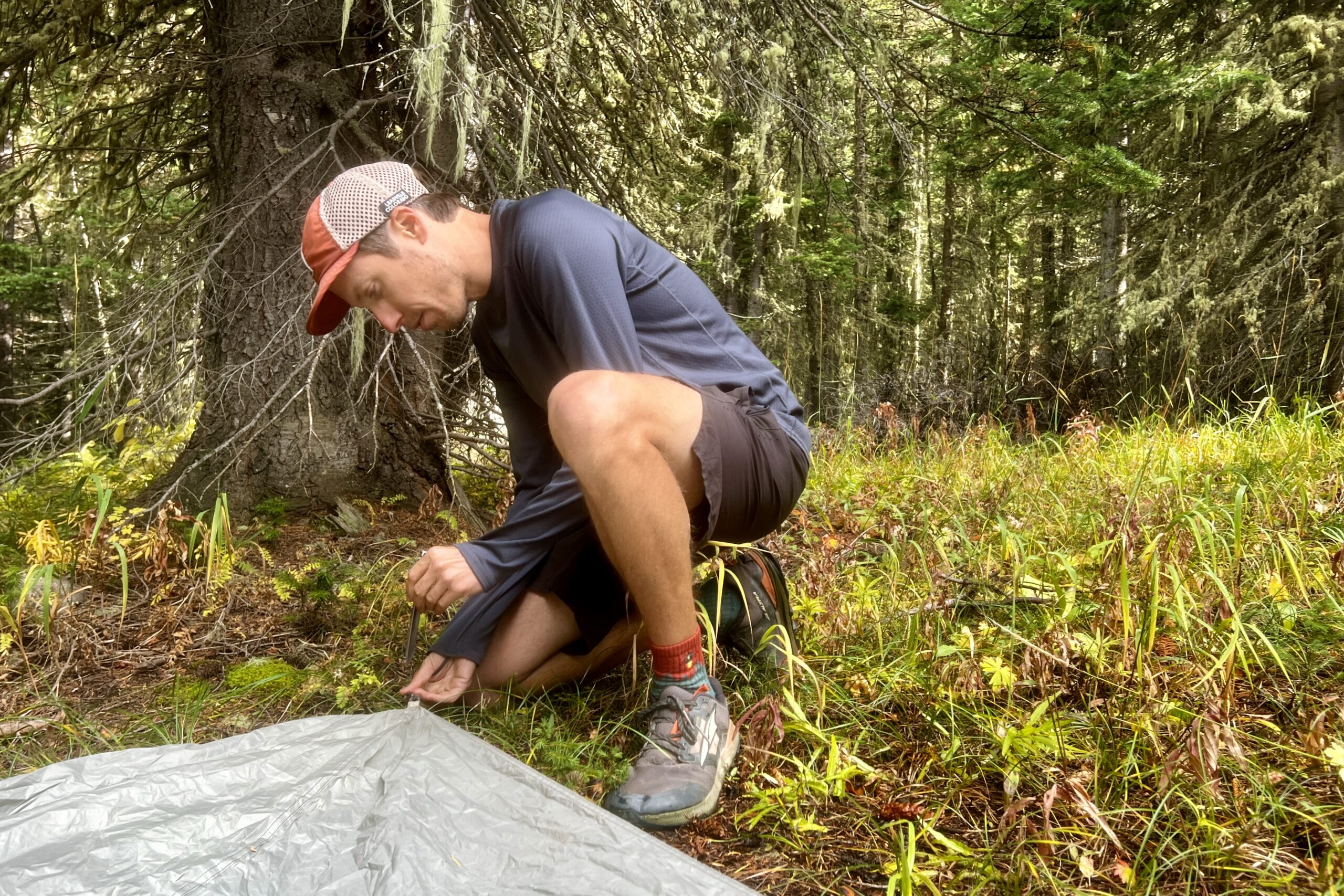 A hiker pounds in a tent stake while wearing the Patagonia Capilene Midweight Crew. Trees are in the background.