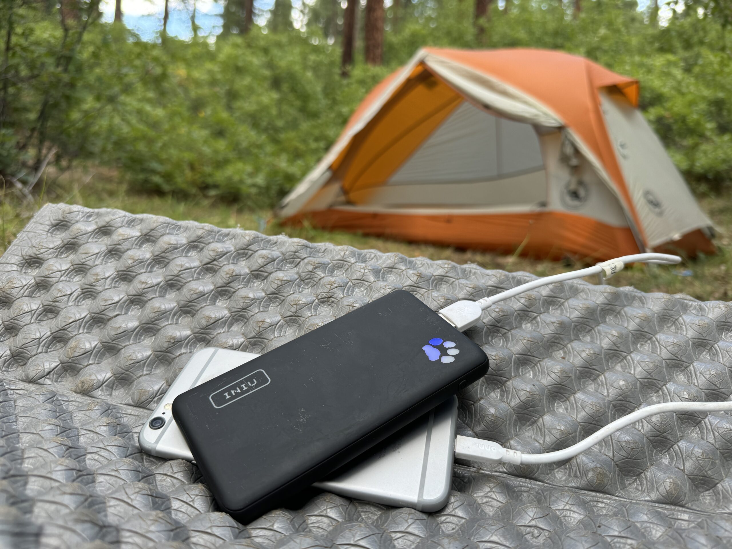 The INIU Megapower 10000 and a phone sit in front of a tent in the woods.