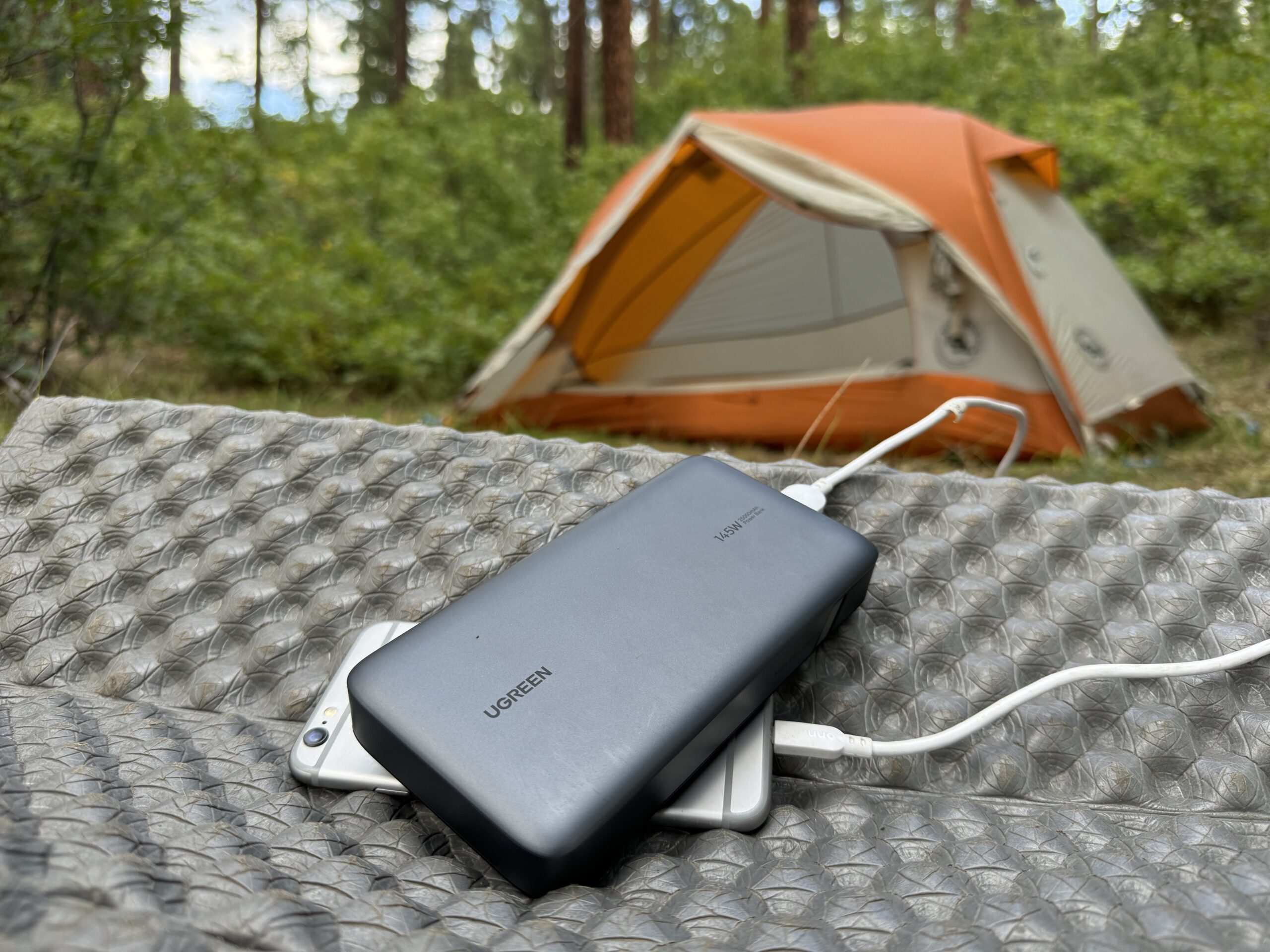 The UGREEN and a phone sit in front of a tent in the woods.