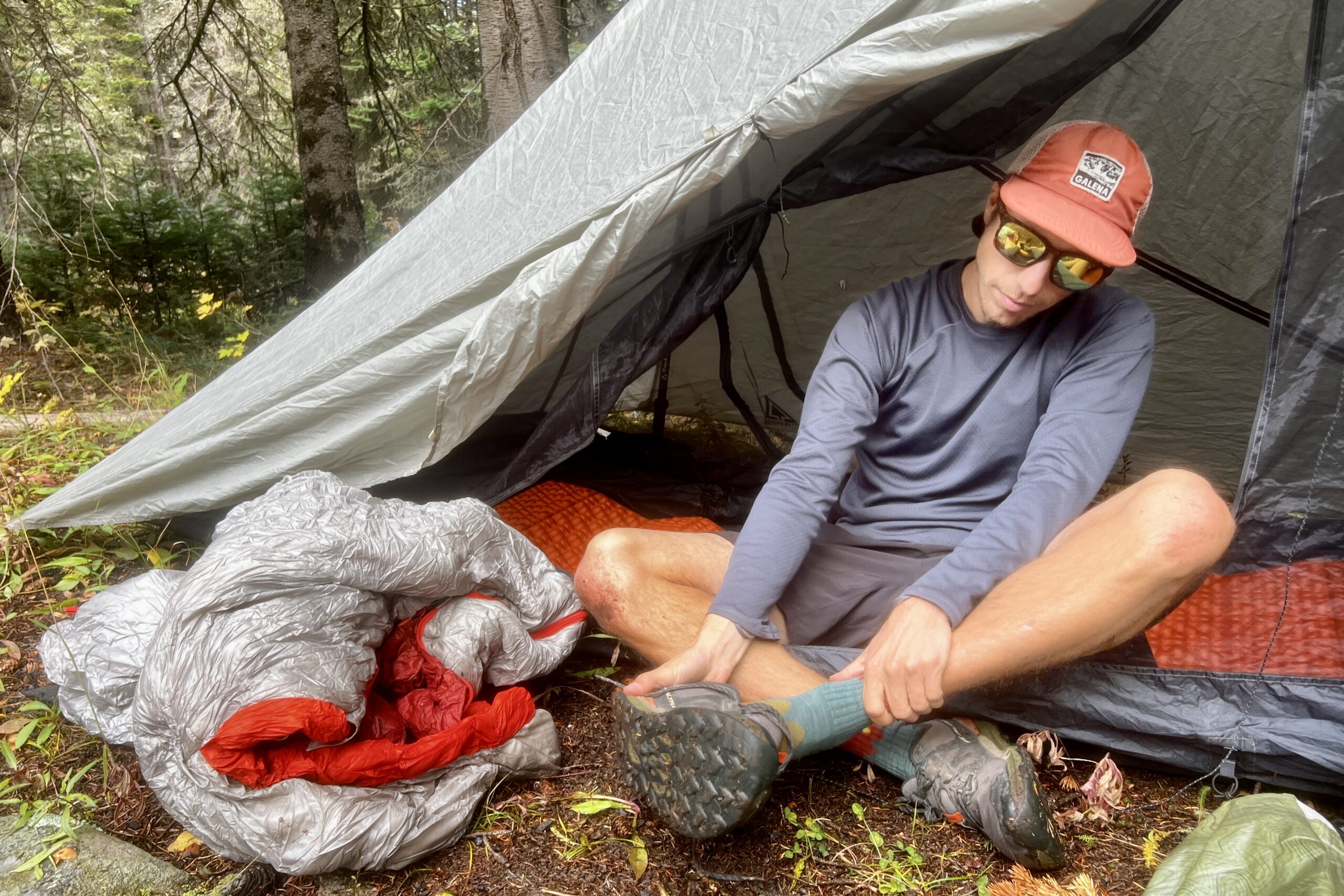 A hiker sitting in his tent and taking off his shoes while wearing the Patagonia Capilene base layer. A tent and trees are in the background with a balled up sleeing bag nearby.