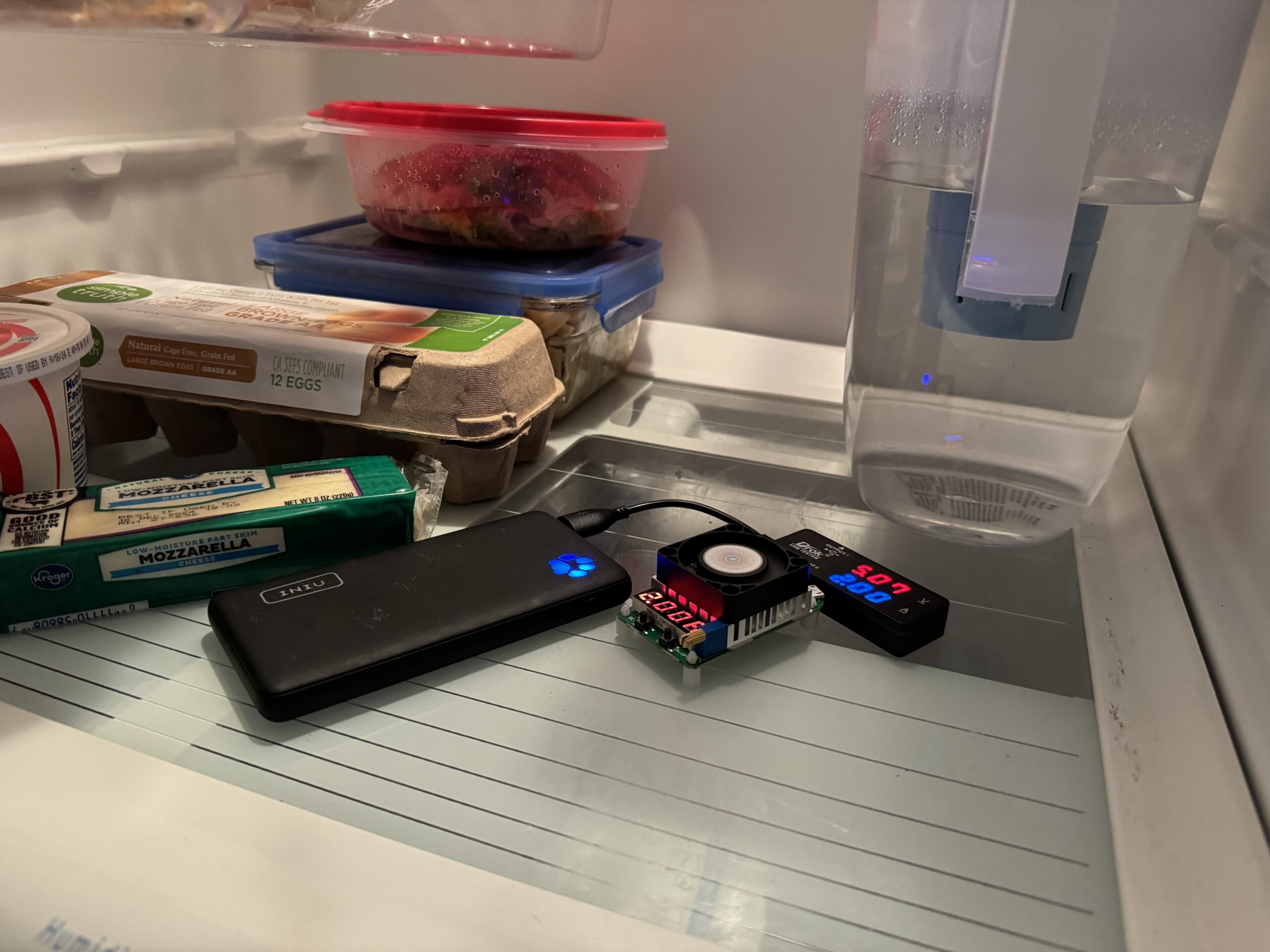 The INIU Megapower 10000 power bank is hooked up to a multimeter and dummy resistor load inside a refrigerator with food.