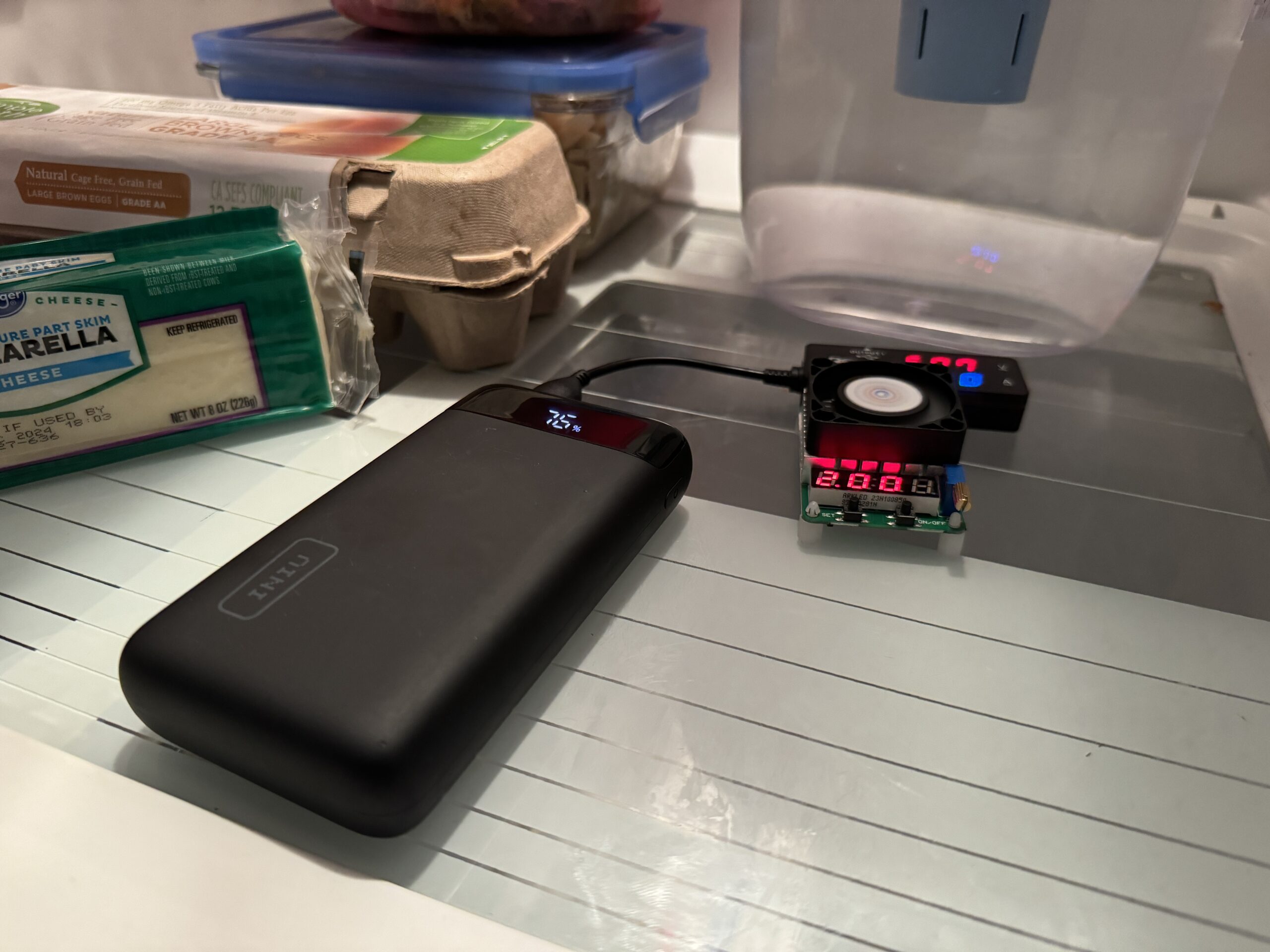 The INIU 20000 PD power bank is hooked up to a multimeter and dummy resistor load inside a refrigerator with food.