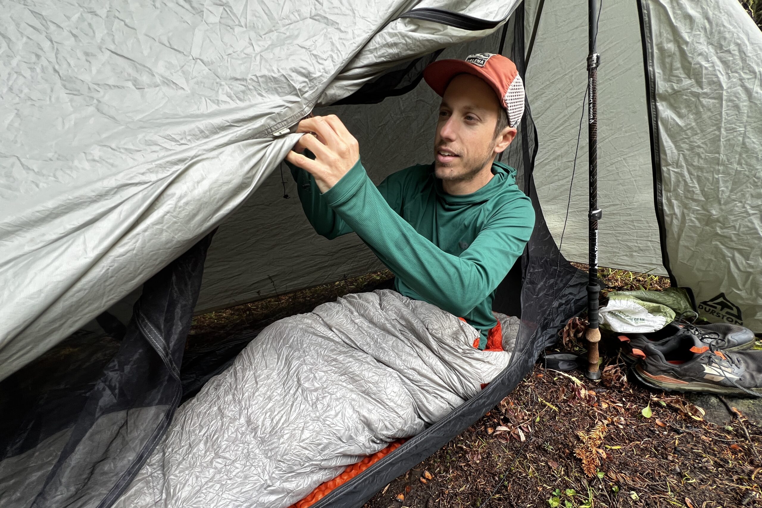 A hiker adjusts his tent while sitting in a sleeping bag, wearing the Outdoor Research Echo Hoodie.