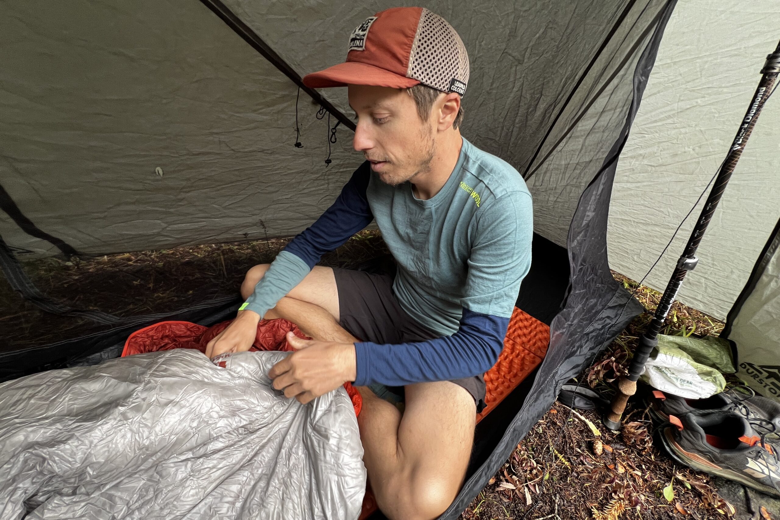 A hiker packing up his sleeping back while wearing the Ortovox Rock'n'Wool base layer. A tent, shoes, and other gear are in the background.