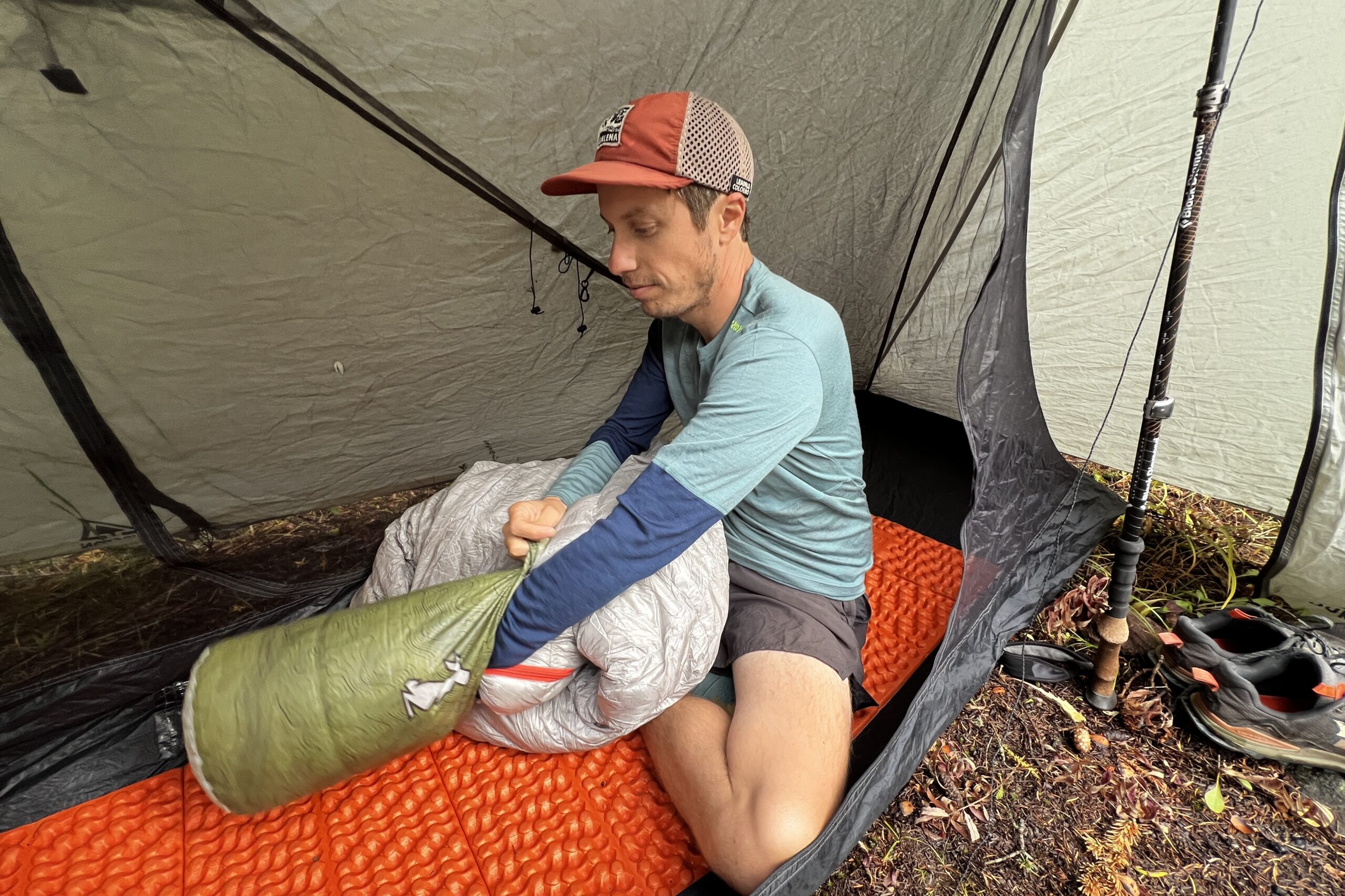 A hiker packing up his sleeping back while wearing the Ortovox Rock'n'Wool base layer. A tent, shoes, and other gear are in the background.