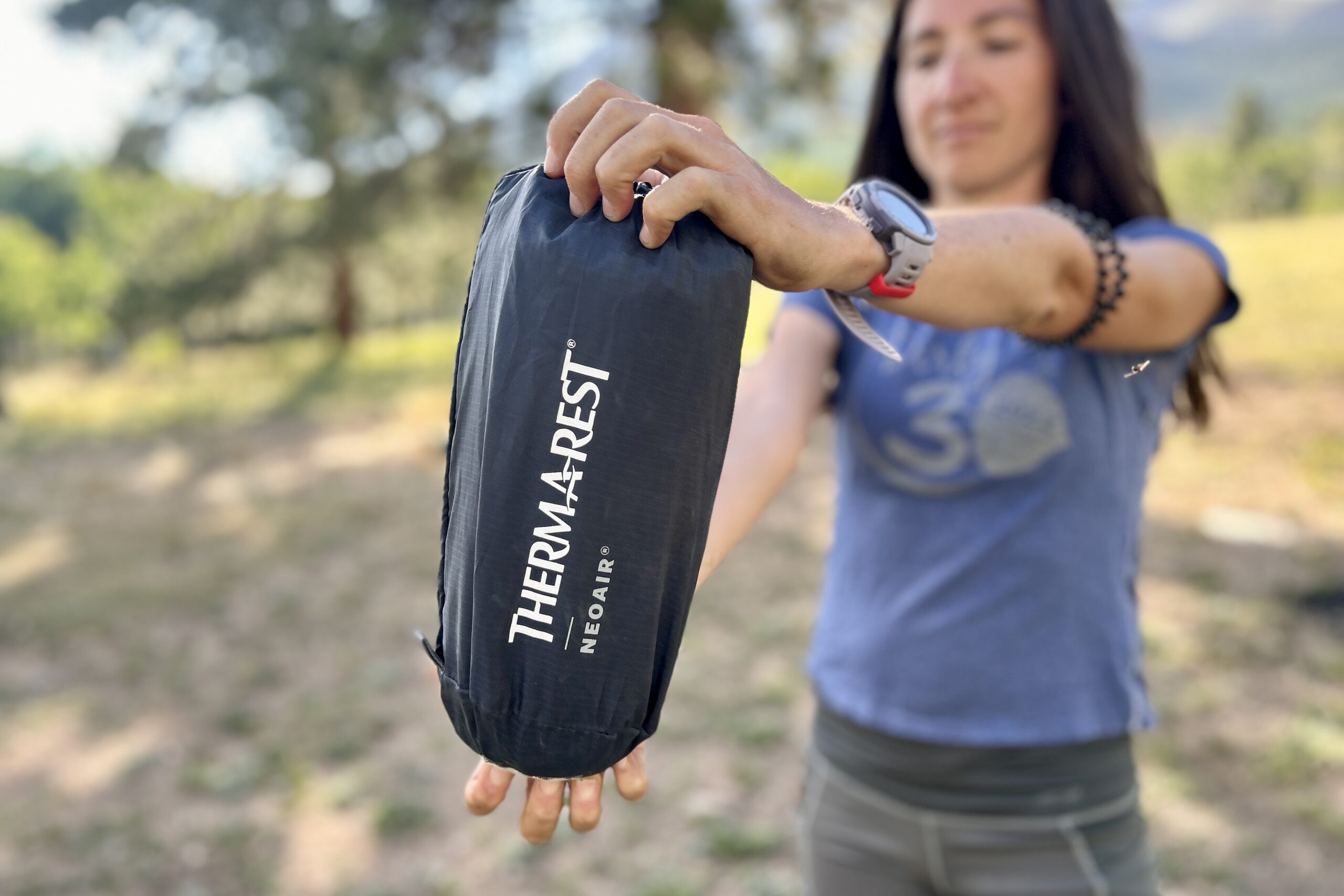 A woman holds a small black stuff sack labeled "Therm-a-Rest NeoAir ".