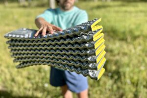 A man stands holding a yellow and grey foam sleeping pad, showcasing the pattern and accordion-style packed size.