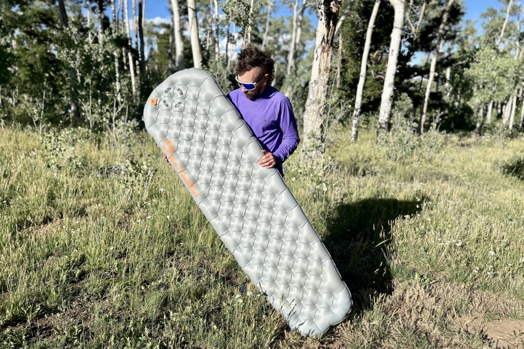 In a large field with trees in the background, a man holds an inflatable grey sleeping pad.