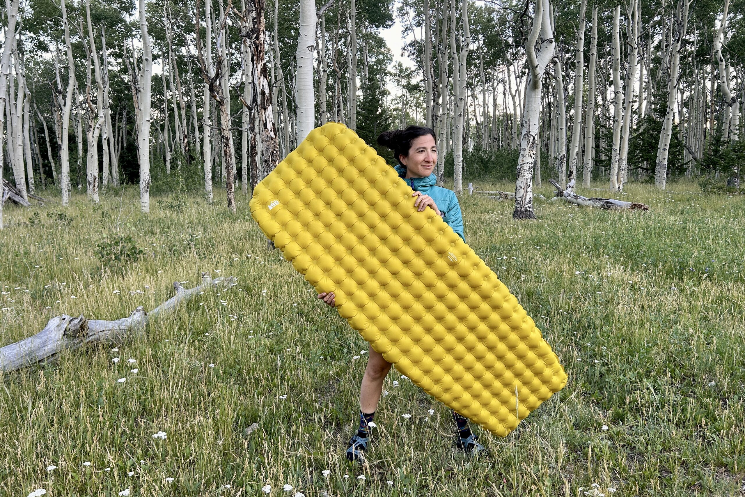 A woman stands next to aspen trees in grass, gripping a large yellow inflatable camping pad.