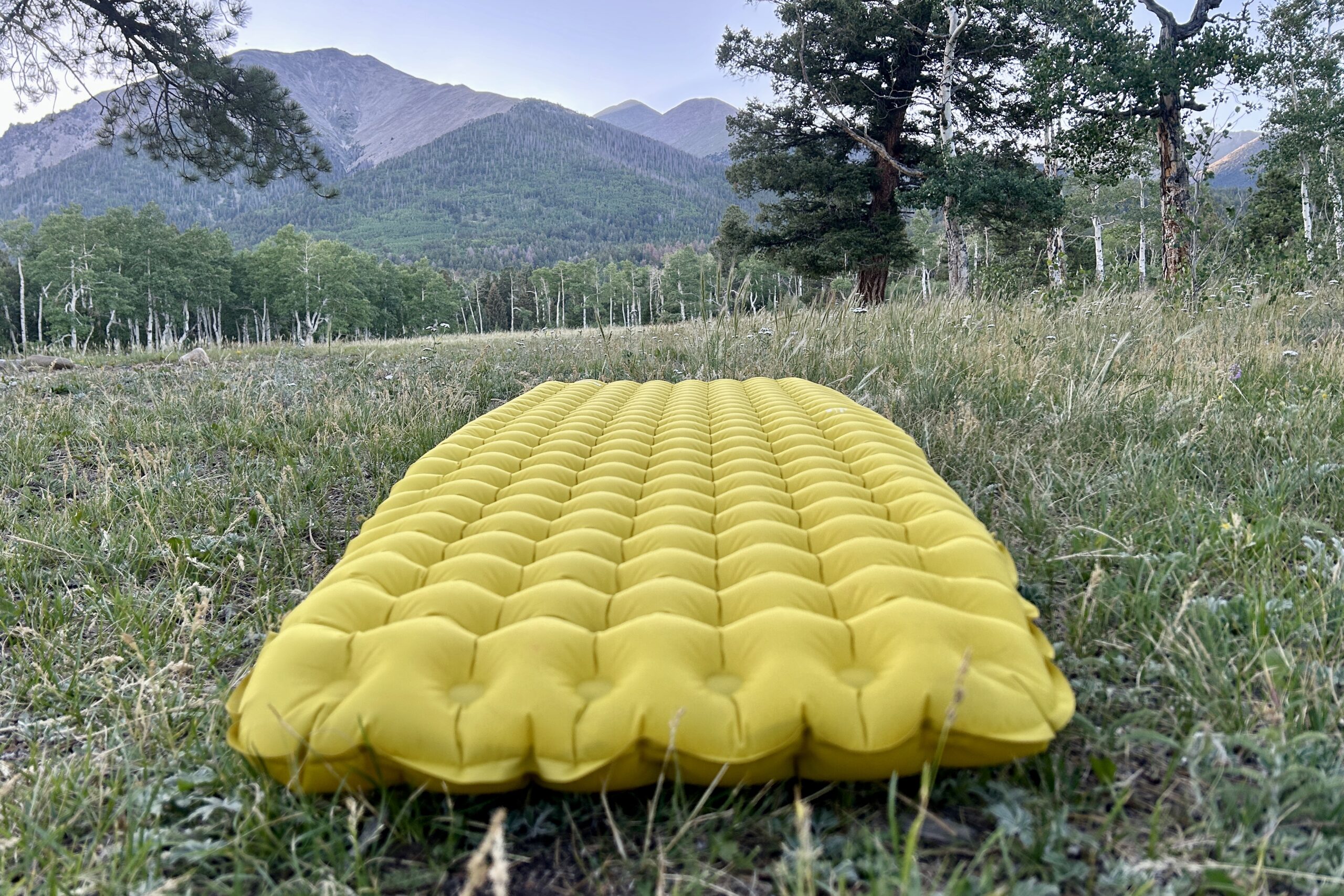 Close up of a yellow inflatable camping pad in the grass with mountains in the background.