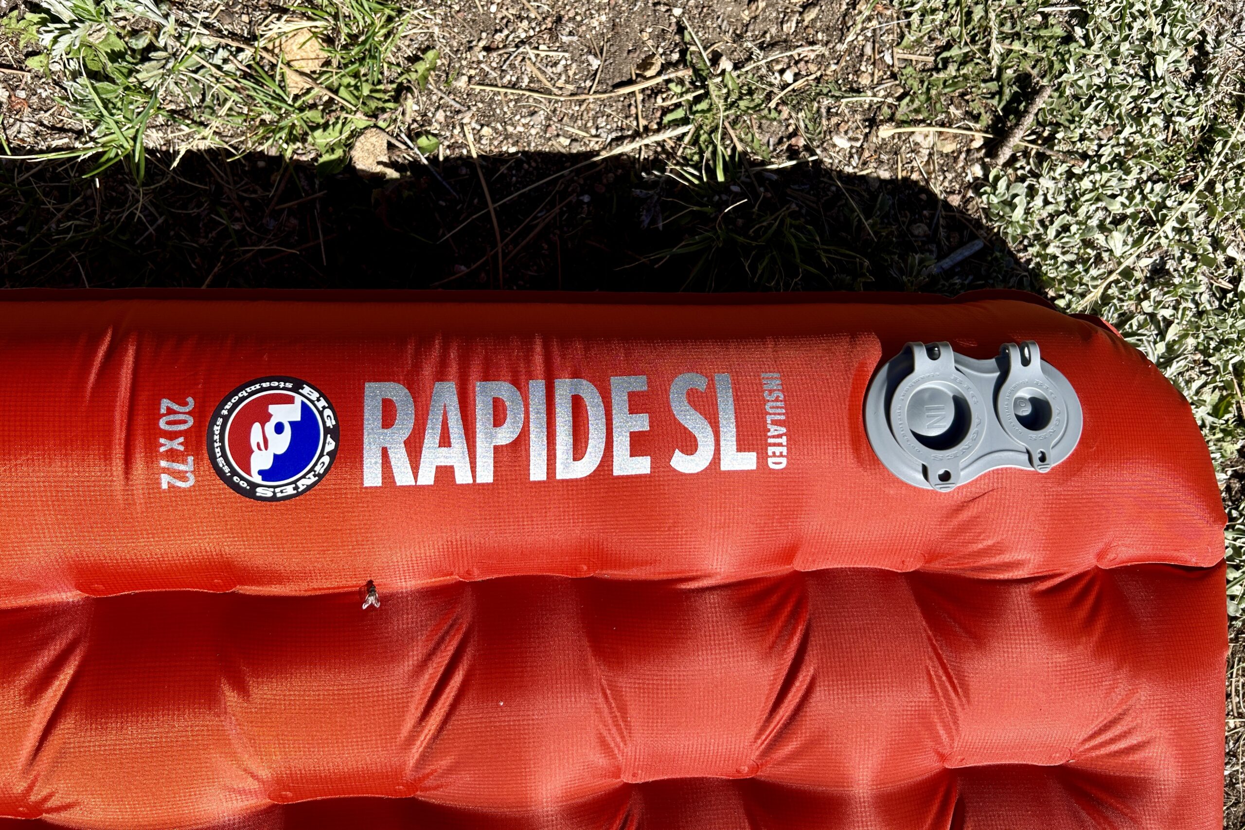 A red inflatable sleeping pad labeled "RAPIDE SL" lays on grass, featuring a Big Agnes logo, and dual air valves, with its quilted bottom visible.