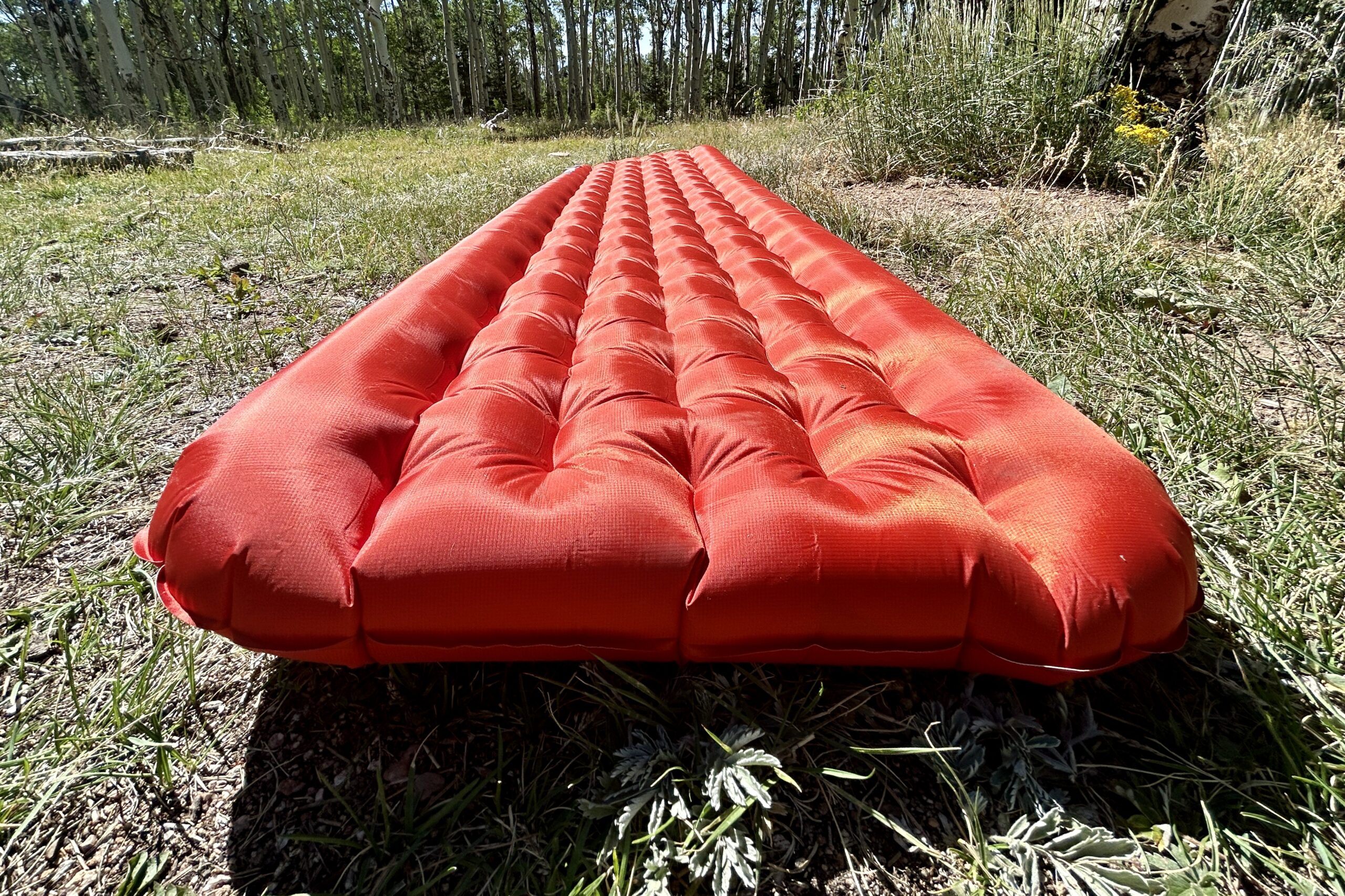 A red inflatable sleeping pad is laying on grass, showcasing its raised side rails and thick cushioning for support.