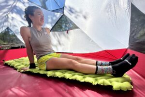 A woman relaxes inside a tent, sitting on a green inflatable sleeping pad. Trees are visible through the tent.