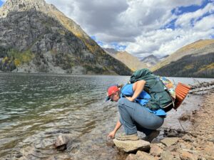 A backpacker wearing the REI Sahara convertible pants kneels next to a lake surrounded by mountains.