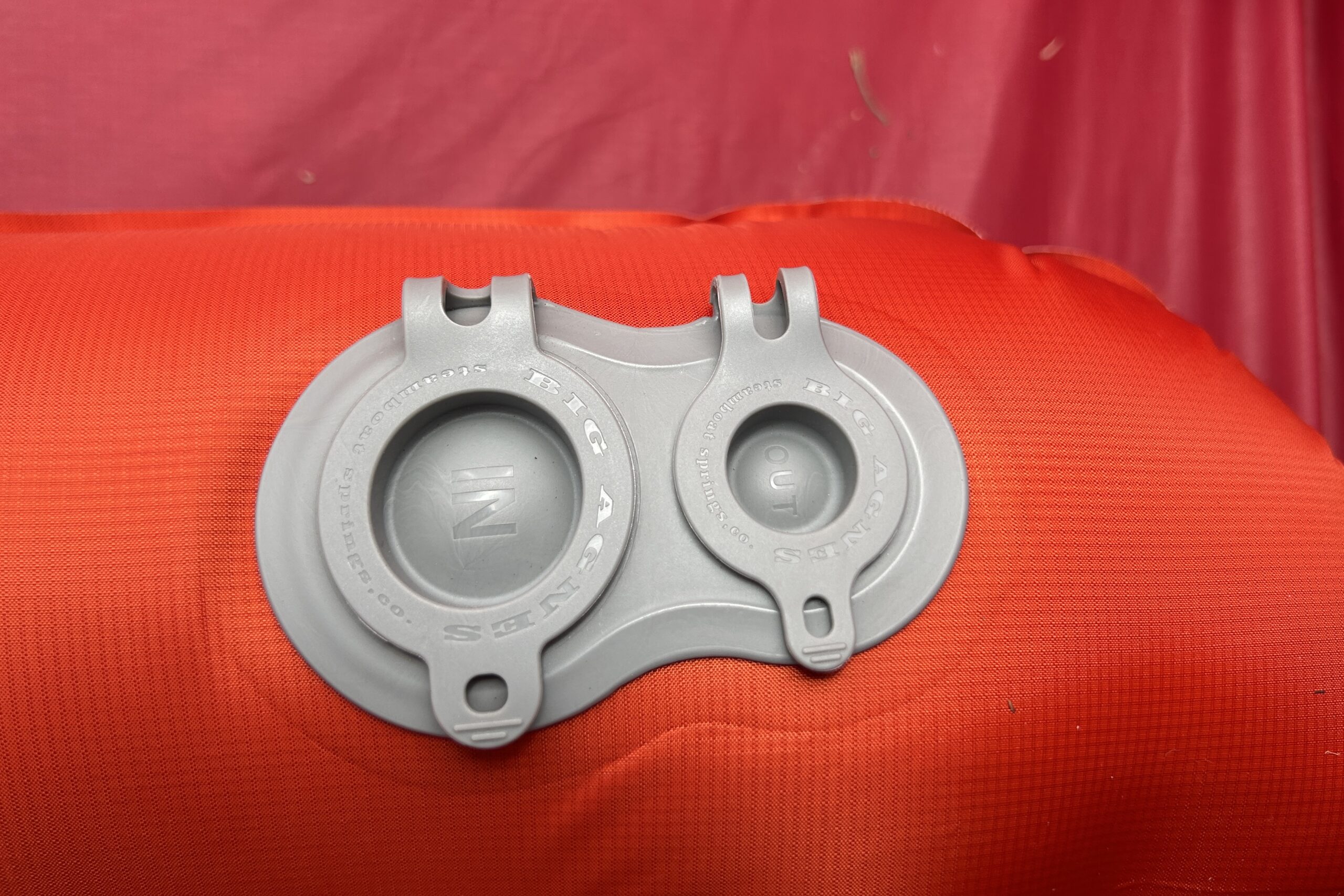 A close-up of a red inflatable sleeping pad with two gray valve caps, one labeled "OUT" and the other "IN", resting on grass.