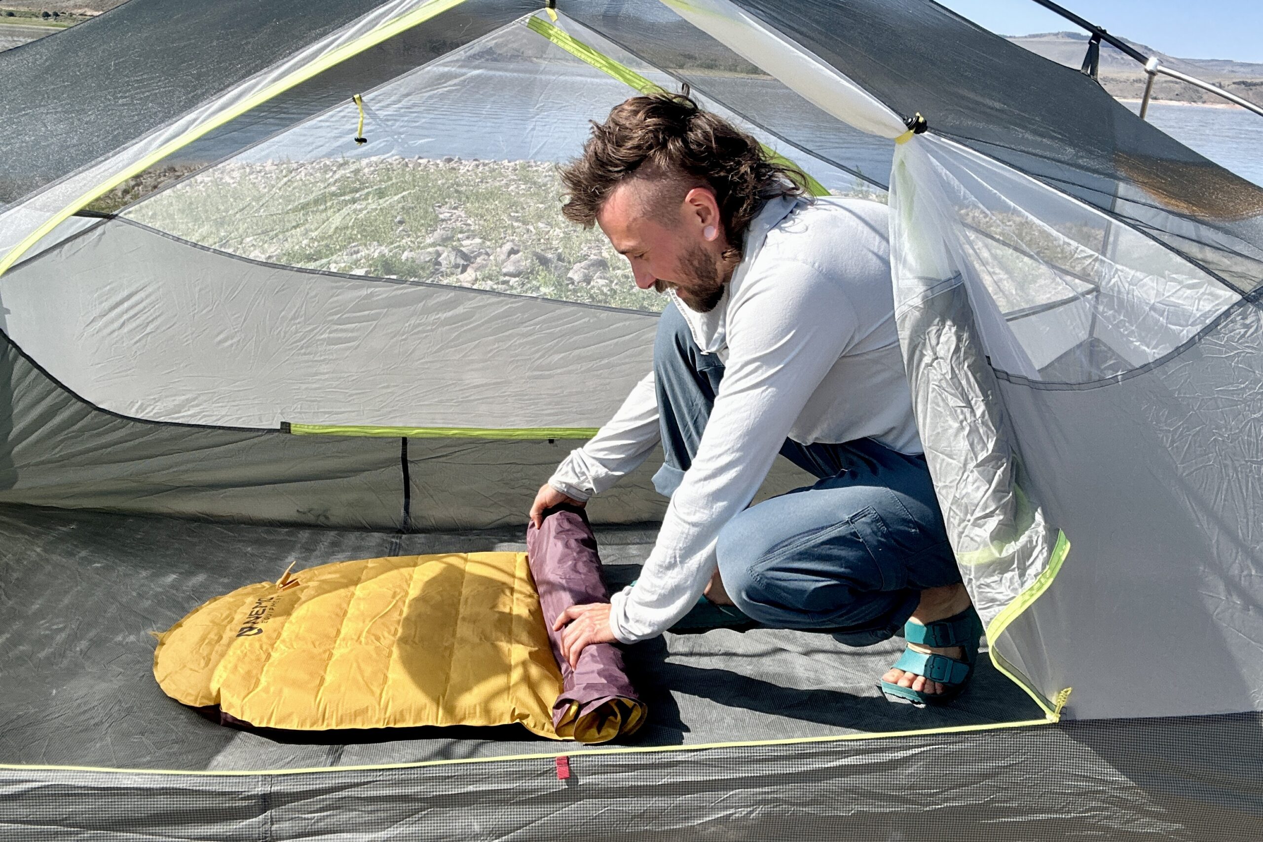 A man rolls up a sleeping pad in a tent with a lake behind him.