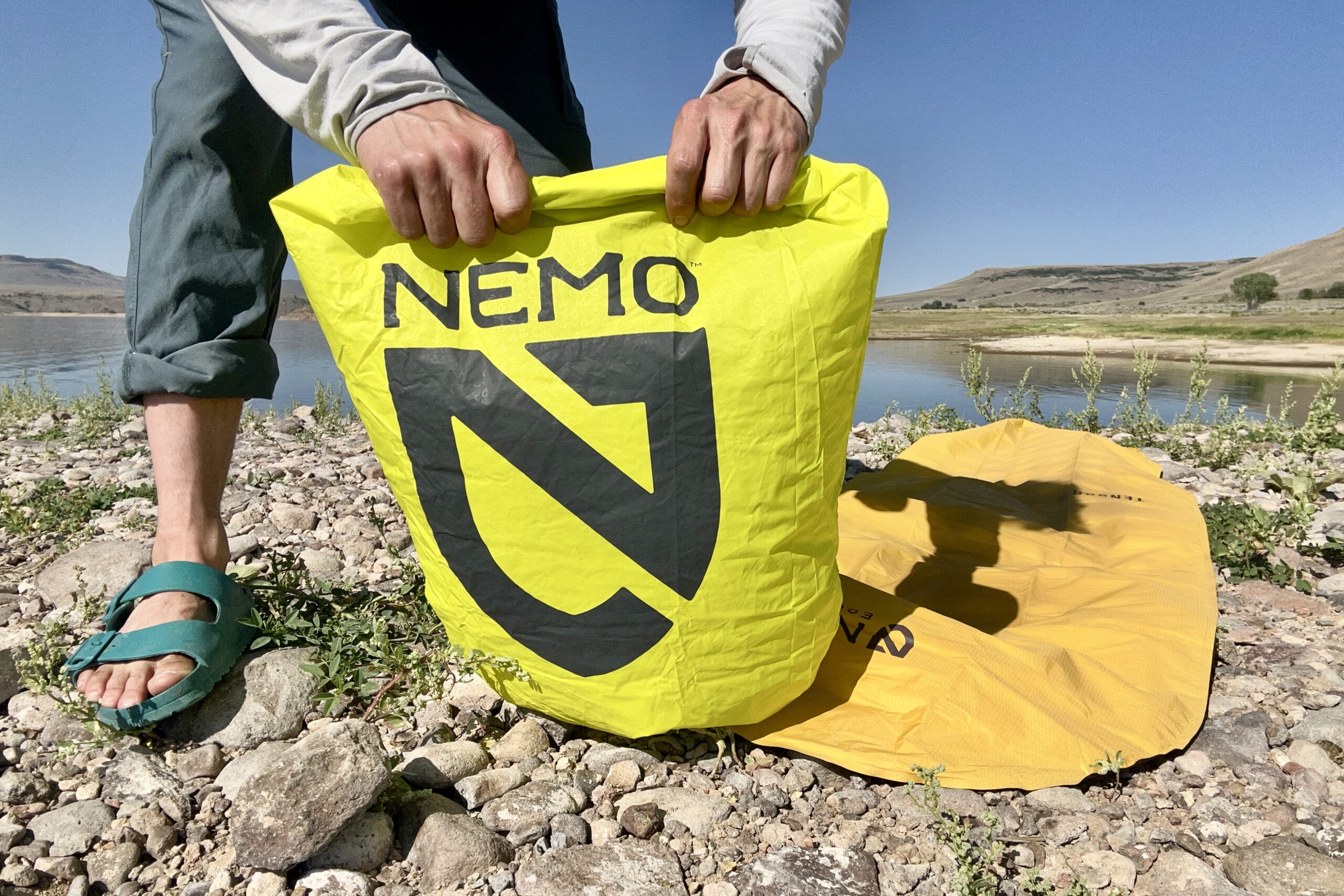 A person uses a bright green pump sack to fill an orange sleeping pad on a lakeside.