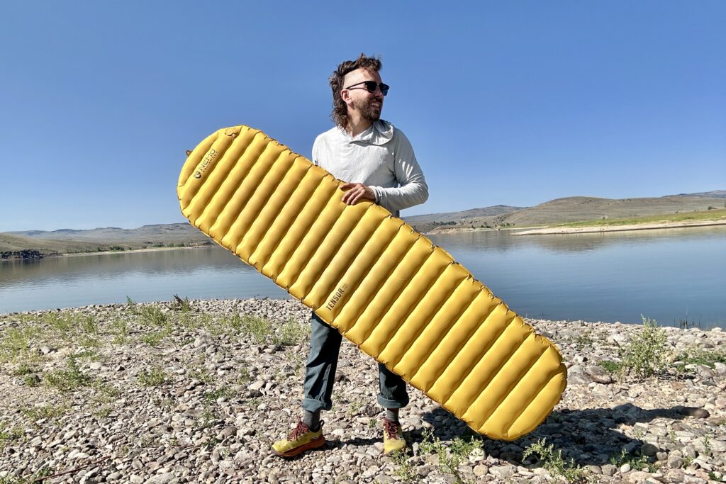 A man stands next to a lake gripping a large yellow inflatable camping pad.