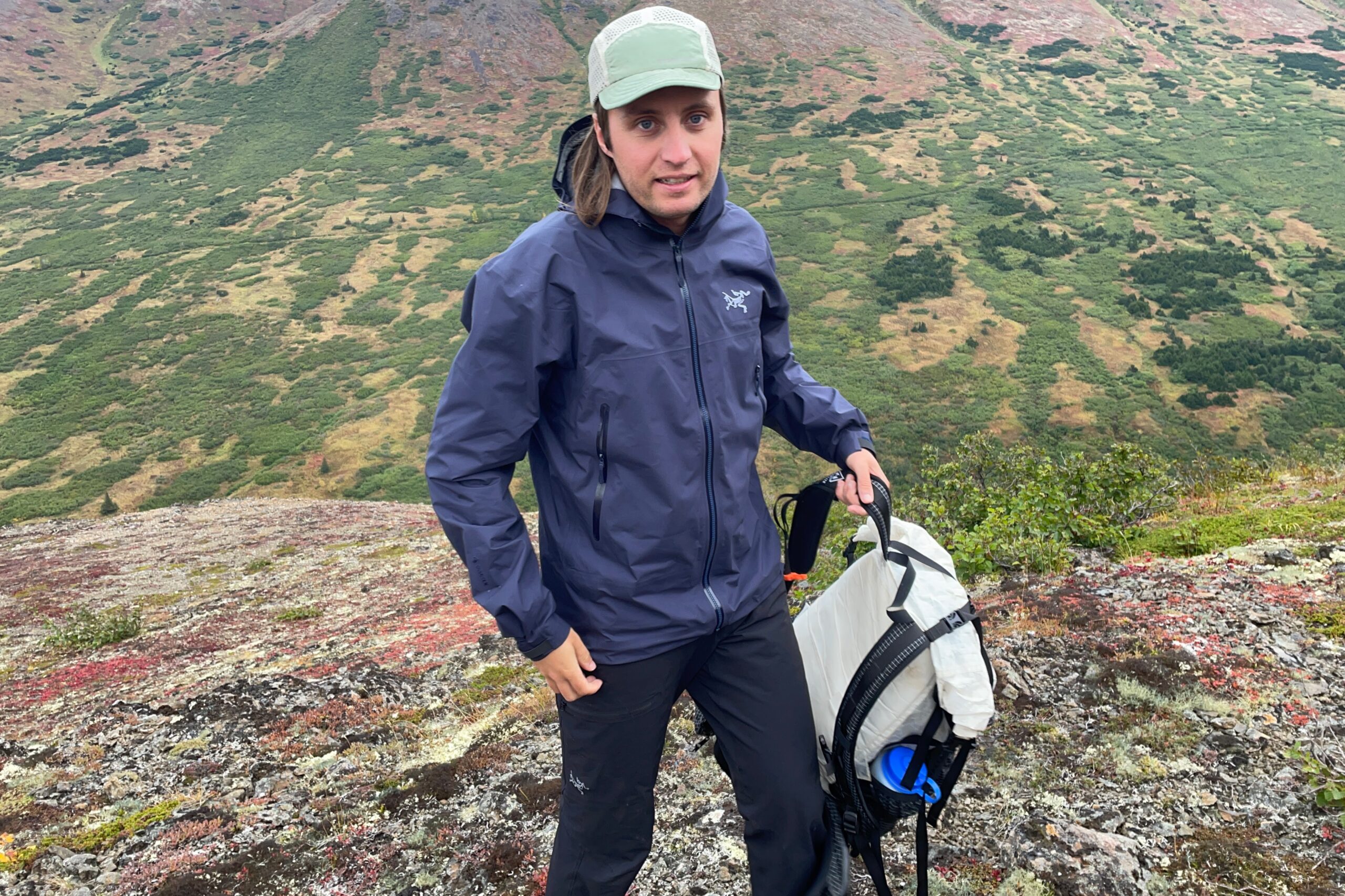 A man holds his backpack while hiking uphill