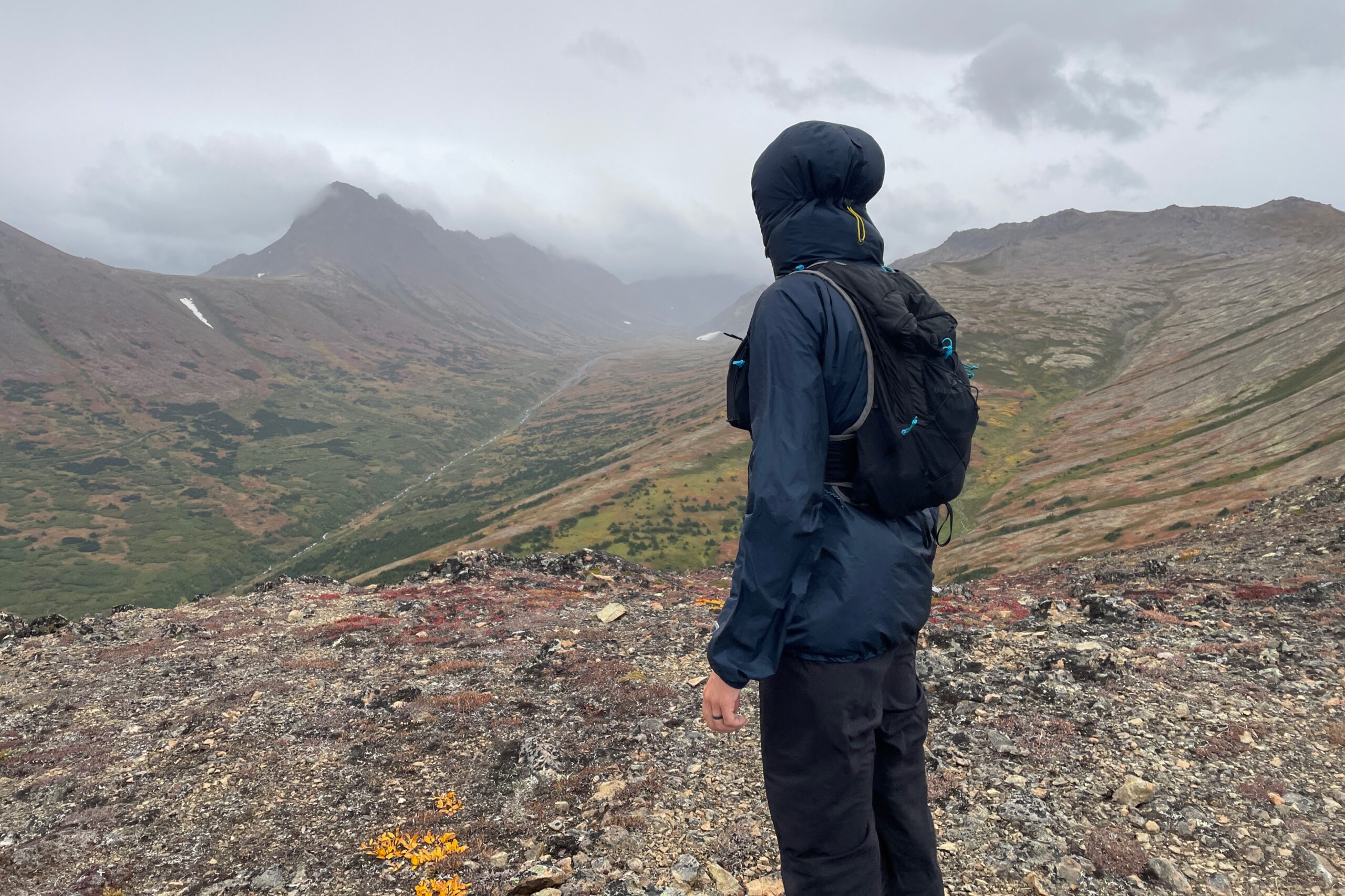 A man admires a mountain view, back towards the camera and hooded against the rain.