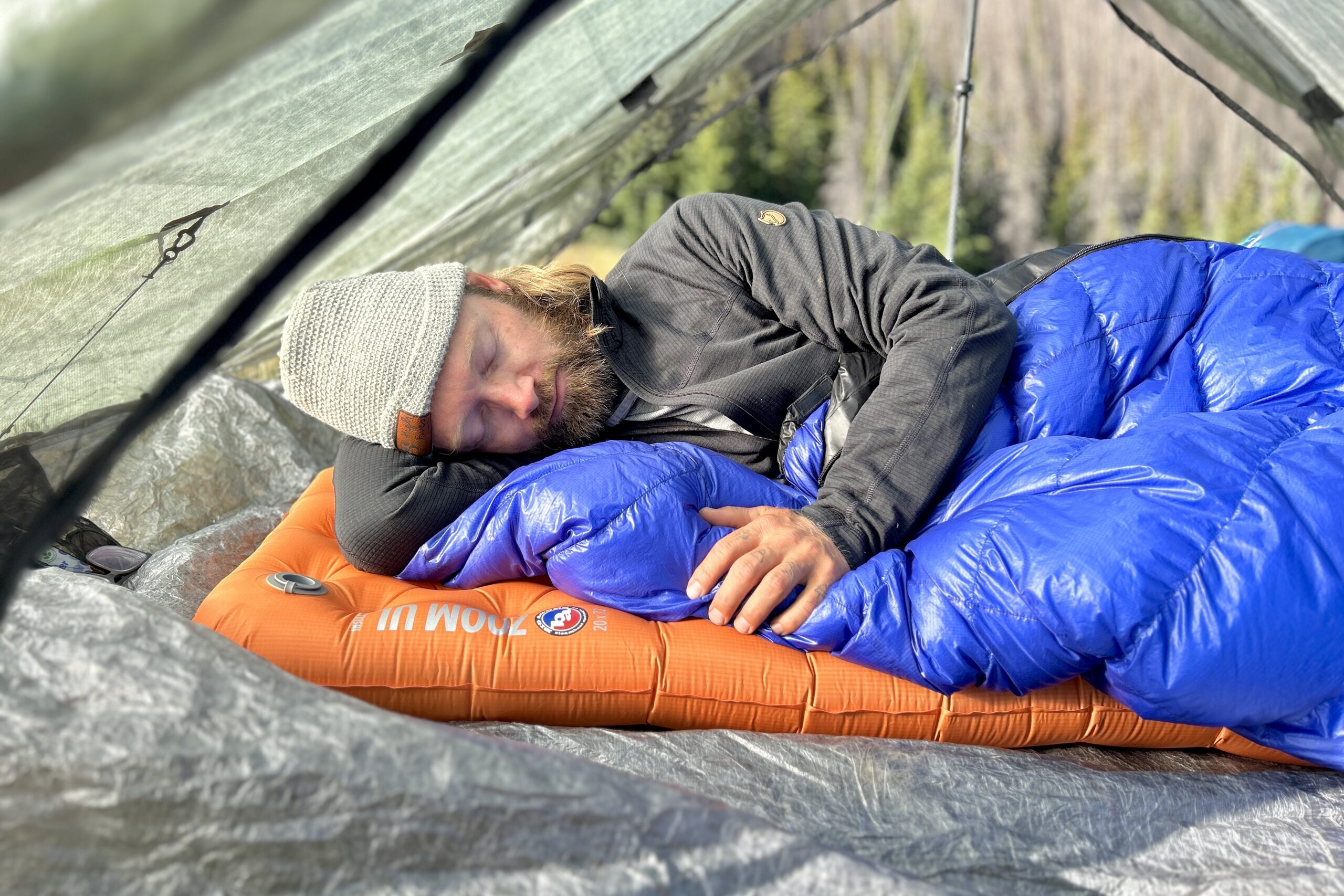 A man peacefully sleeps in a vibrant orange and blue sleeping bag in a tent with forest in background.