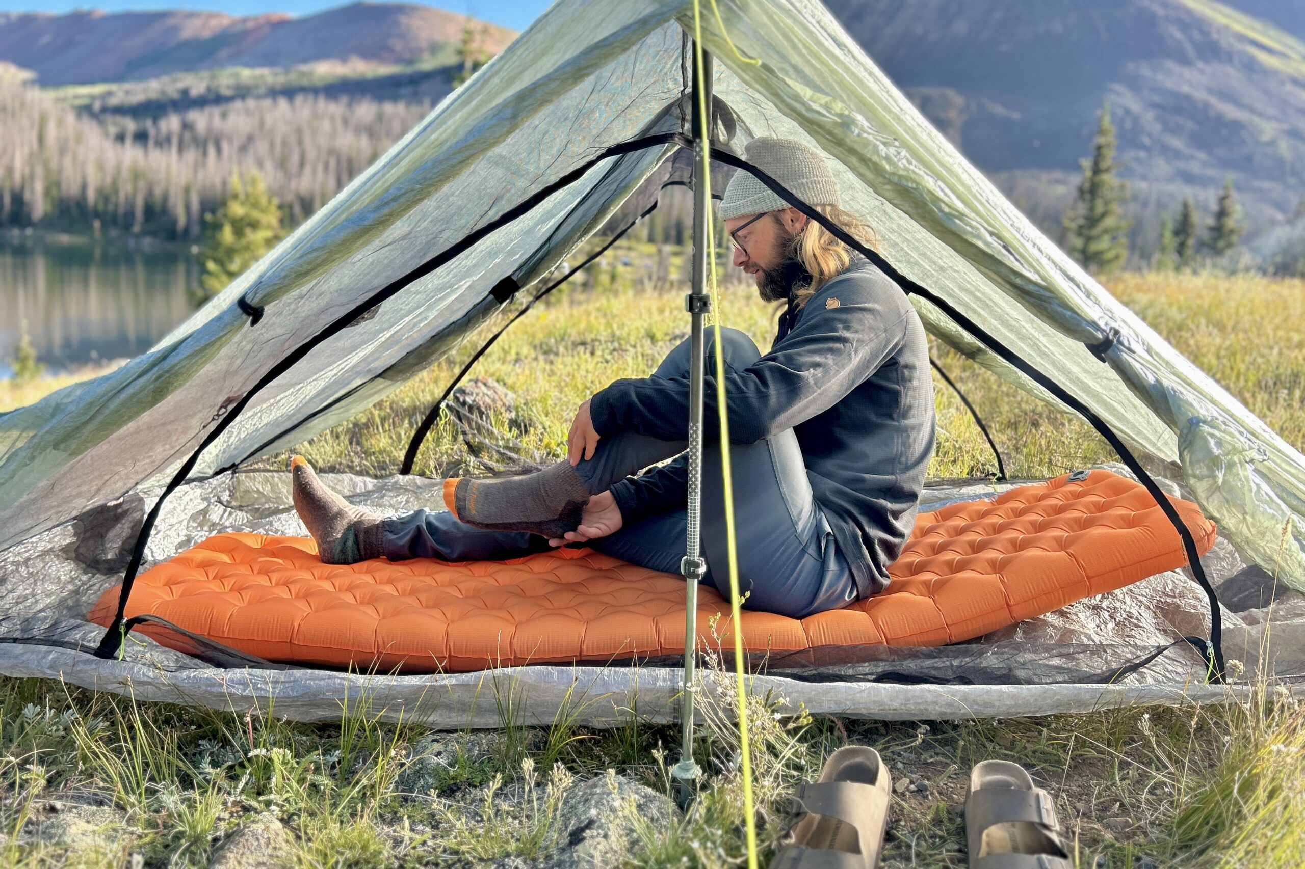 A man sits inside a tent on an orange mat, surrounded by camping gear and mountains.