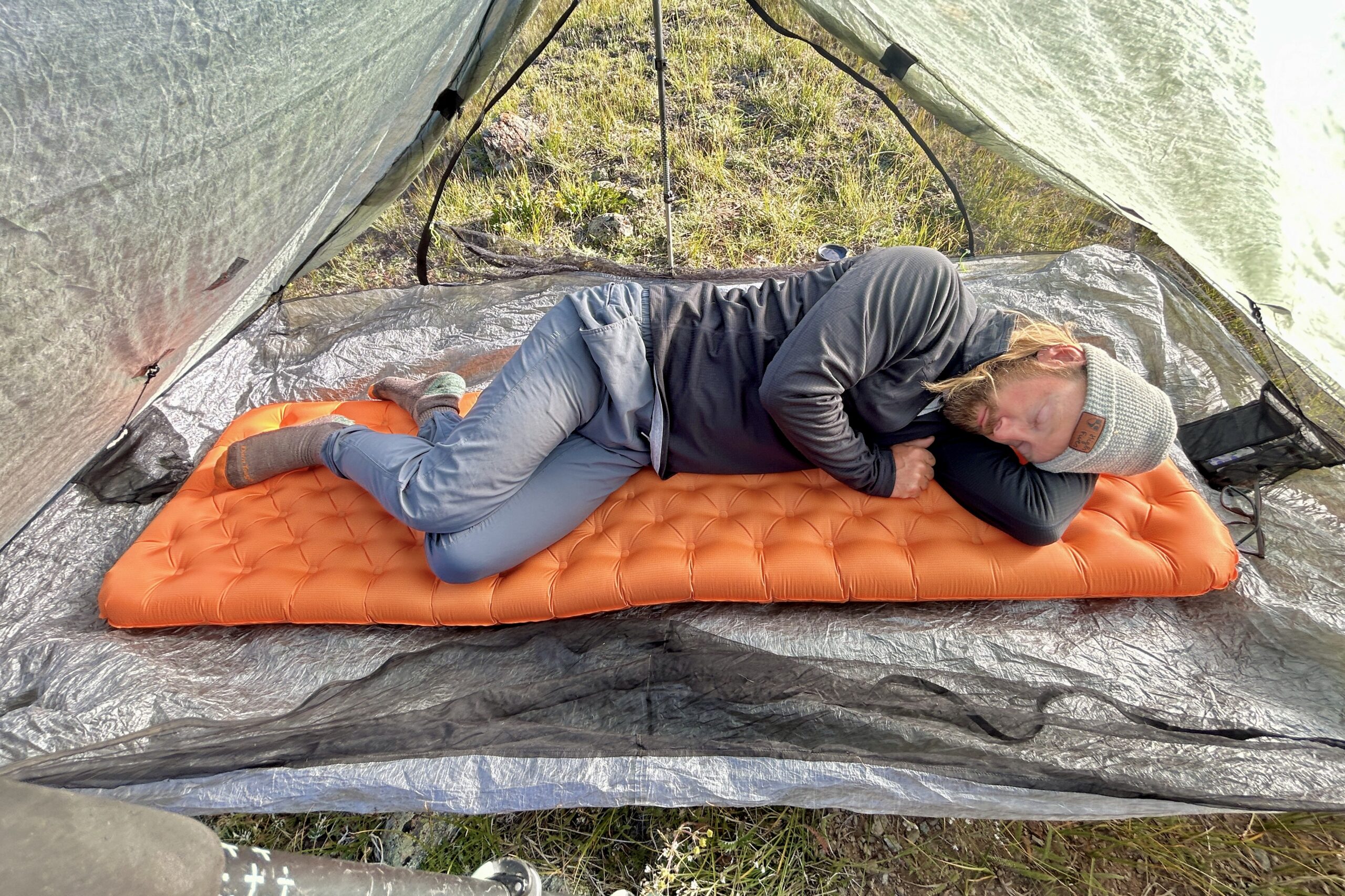 A man rests comfortably on an orange sleeping pad in a tent in a field of green grass.