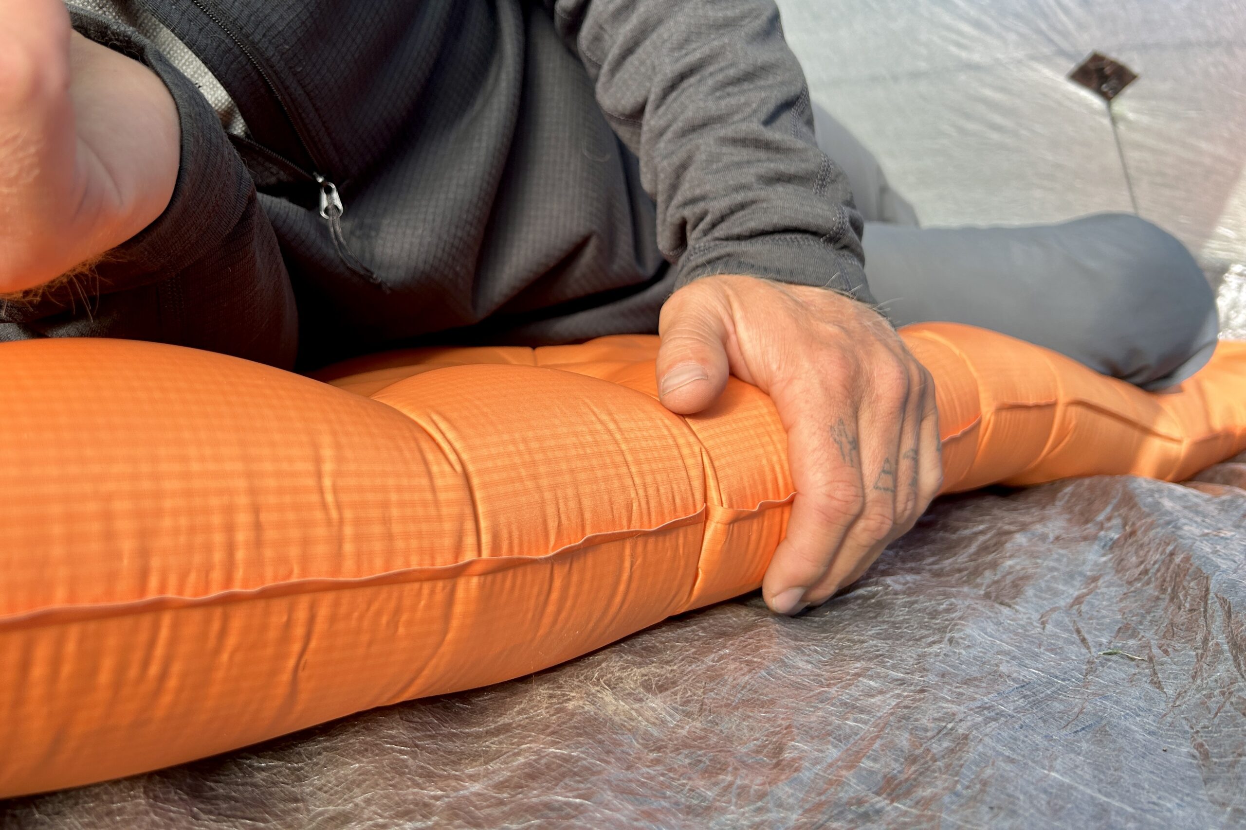 A person lays on an orange sleeping pad with his hand on the side to show its thickness.