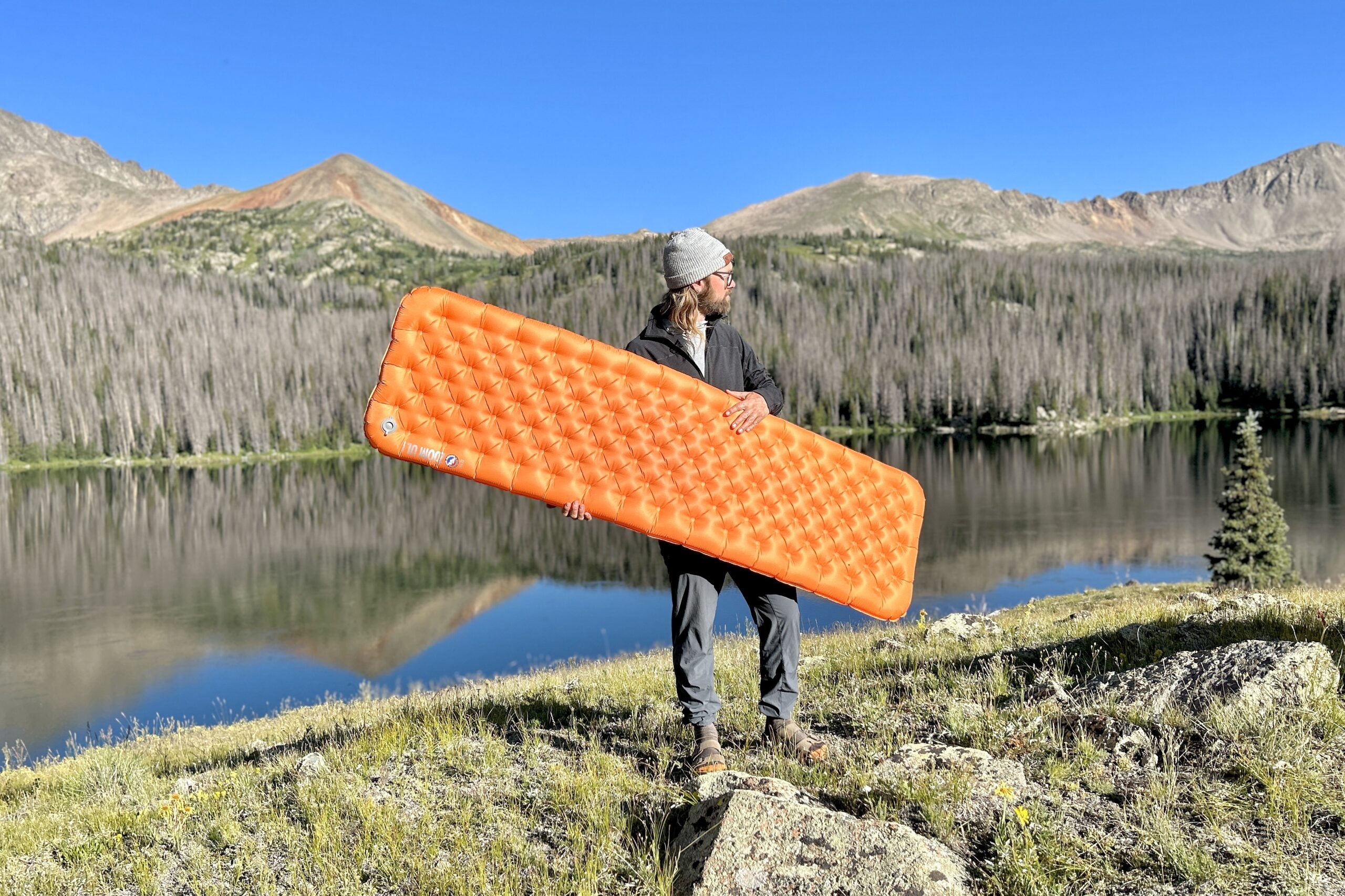 A man stands in front of a lake and mountains holding an orange inflatable mattress.