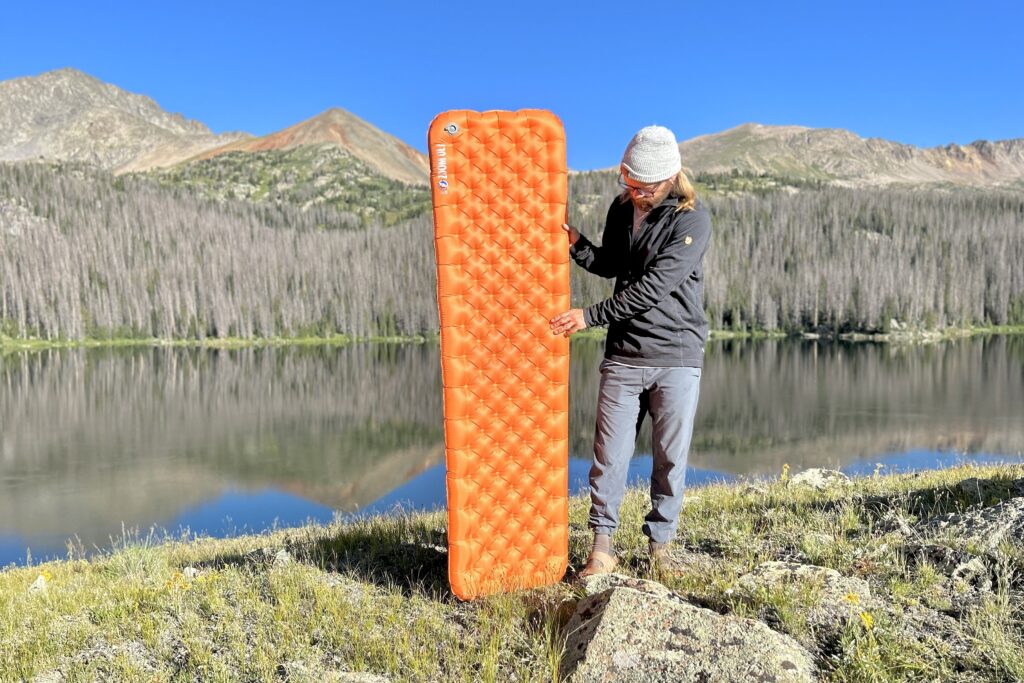 A man stands in front of a lake and mountains holding an orange inflatable mattress.