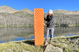 A man stands in front of a lake and mountains holding an orange inflatable mattress.