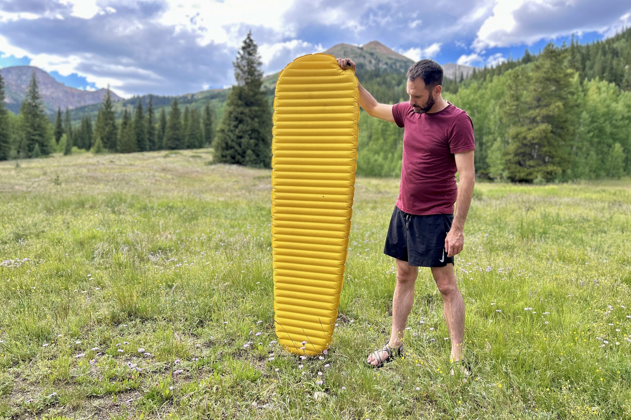 A man stands outdoors, holding a large yellow sleeping pad, with forest and mountains in the background.