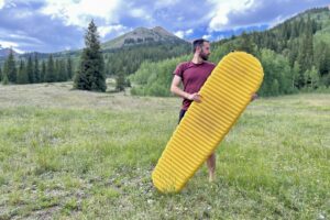 A man stands outdoors, holding a large yellow sleeping pad, with forest and mountains in the background.