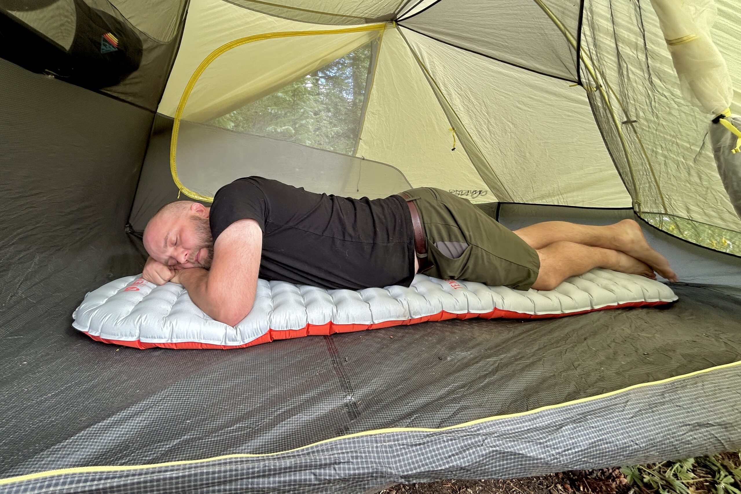 Inside a tent, a bald man lays on his stomach on an an inflatable pad.