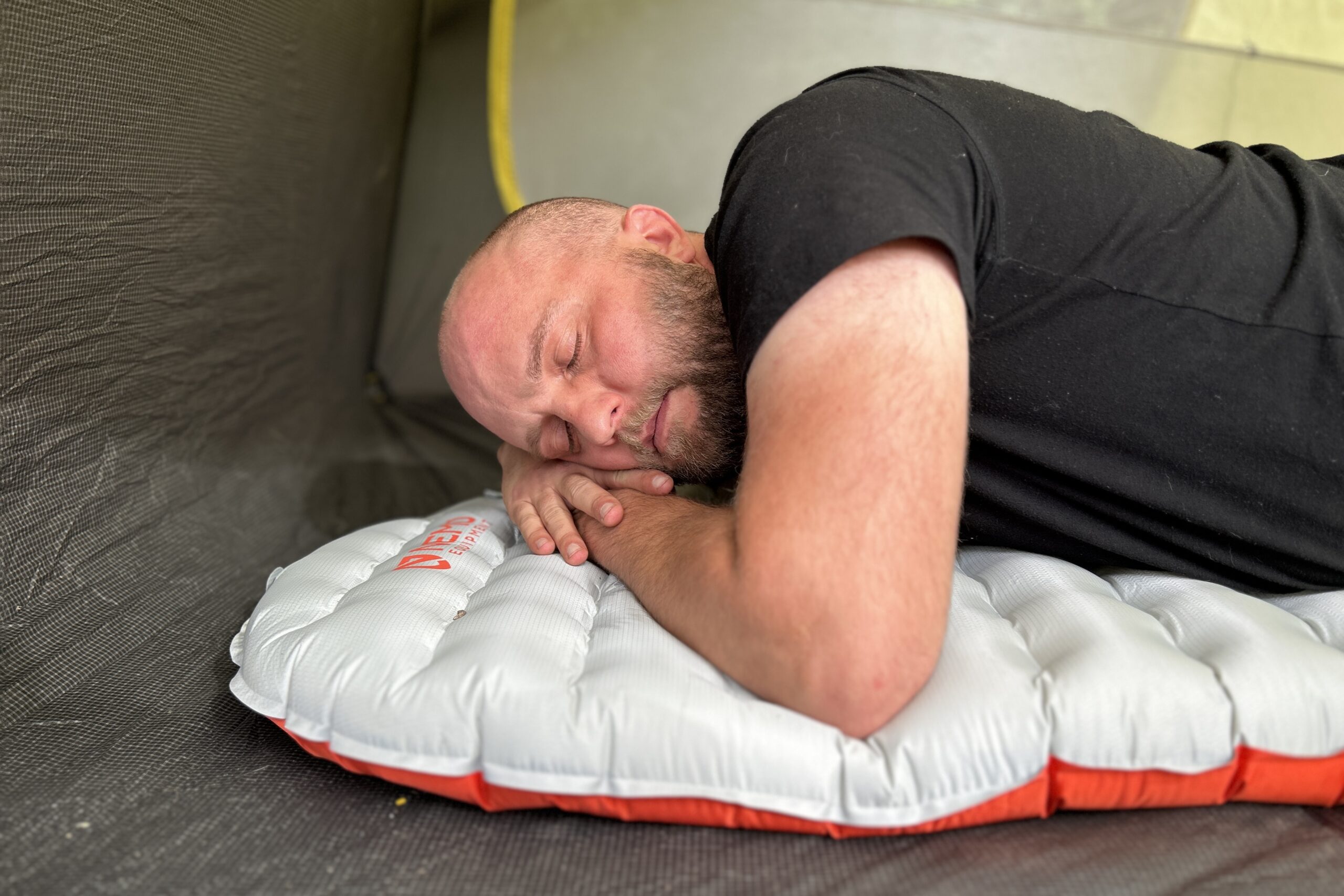 Inside a tent, a bald man lays on his stomach with hands under his head on an inflatable pad.